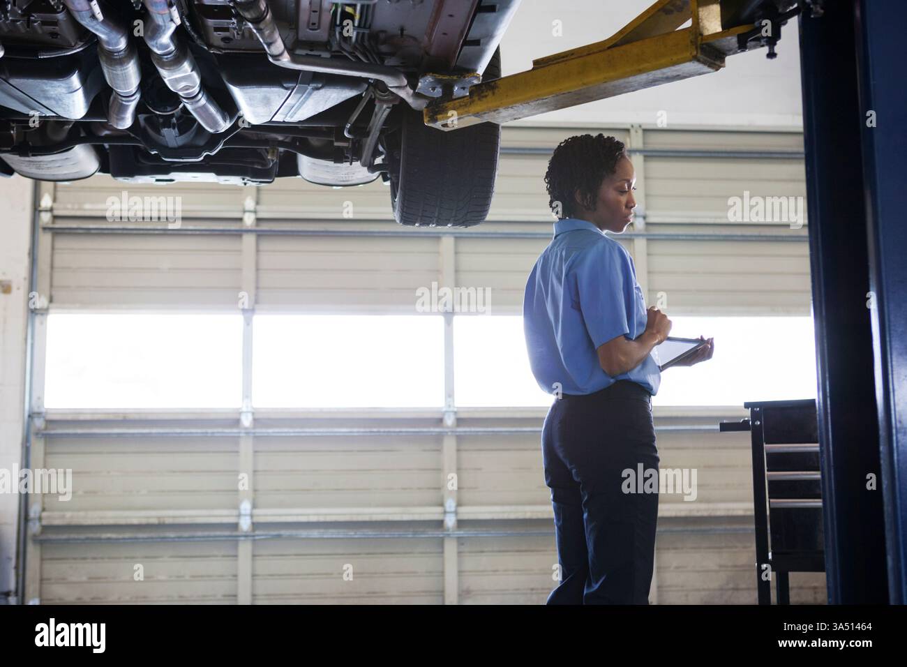 Schwarze Mechanikerin steht neben einem Auto auf einem Aufzug und hält ein Tablet. In der Autowerkstatt wird auf Fachwissen und digitalen Workflow geachtet. Ein professionelles, technisches Image, ideal für den Automobildienst und Frauen im Handwerk. Stockfoto