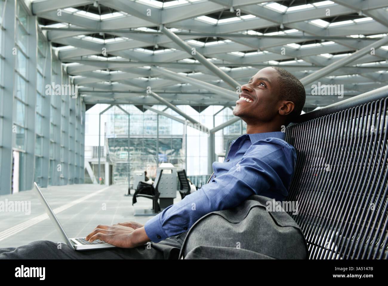 Porträt eines fröhlichen jungen Geschäftsmannes mit Laptop an einem Bahnhofsterminal. Dieses moderne, von Reisen inspirierte Bild zeigt einen lächelnden Profi bei der Arbeit, während er in einem Transitknoten wartet, perfekt für Geschäftsreisen und Arbeitsplatzthemen. Stockfoto