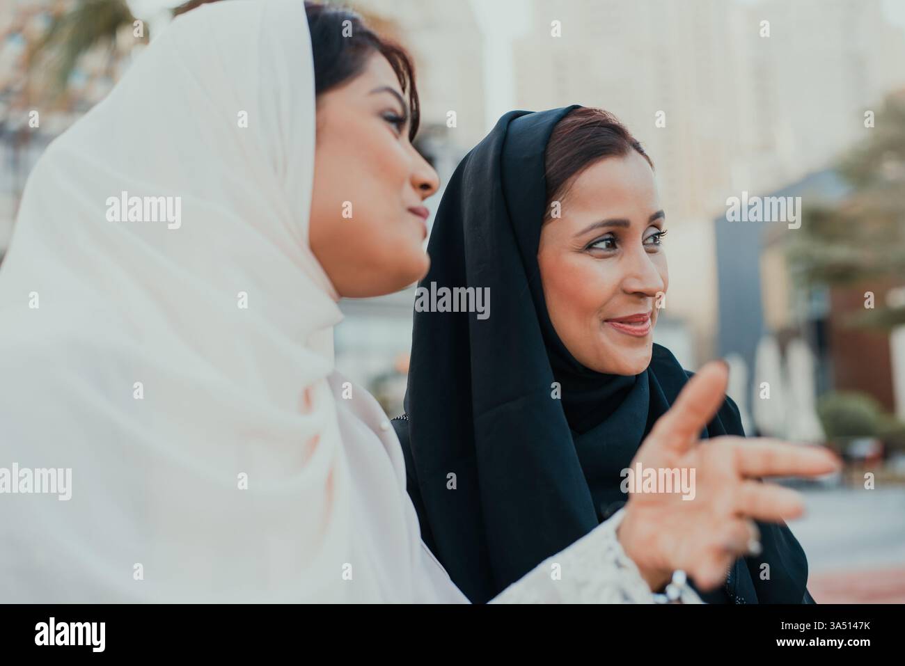 Indische Frauen, die Abaya tragen, stehen tagsüber auf einer Stadtstraße, schauen weg und reden. Die urbane Umgebung vermittelt zeitgenössische bescheidene Mode und soziale Verbindung. Geeignet für Mode-, Reise- und Lifestyle-Kampagnen im Nahen Osten und Südasien. Stockfoto