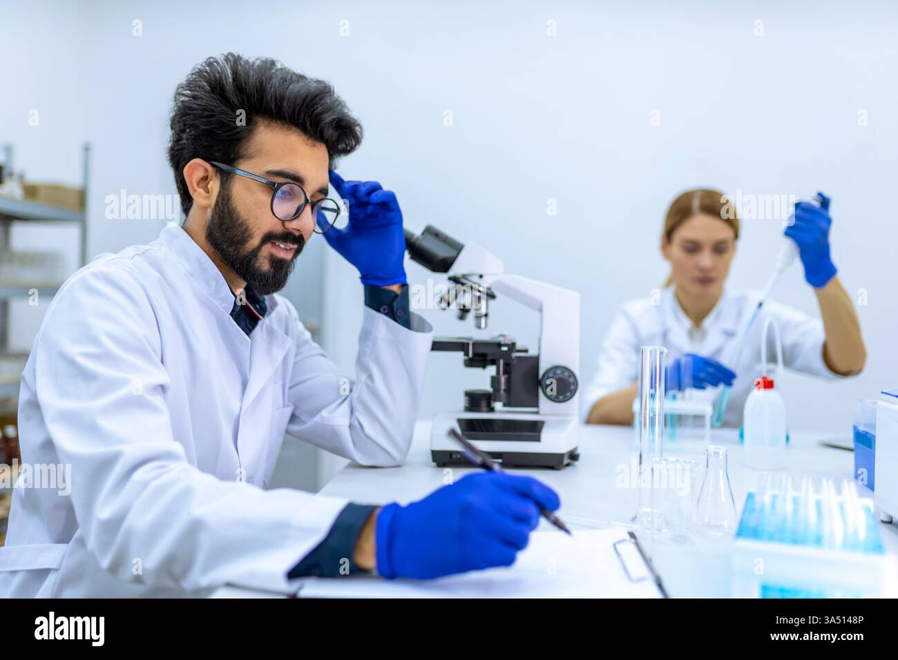 Ein lächelnder indischer Wissenschaftler macht Notizen von seiner Untersuchung, während er am Tisch neben einer kaukasischen Kollegin sitzt, die an Flüssigchemikalien arbeitet ... Stockfoto