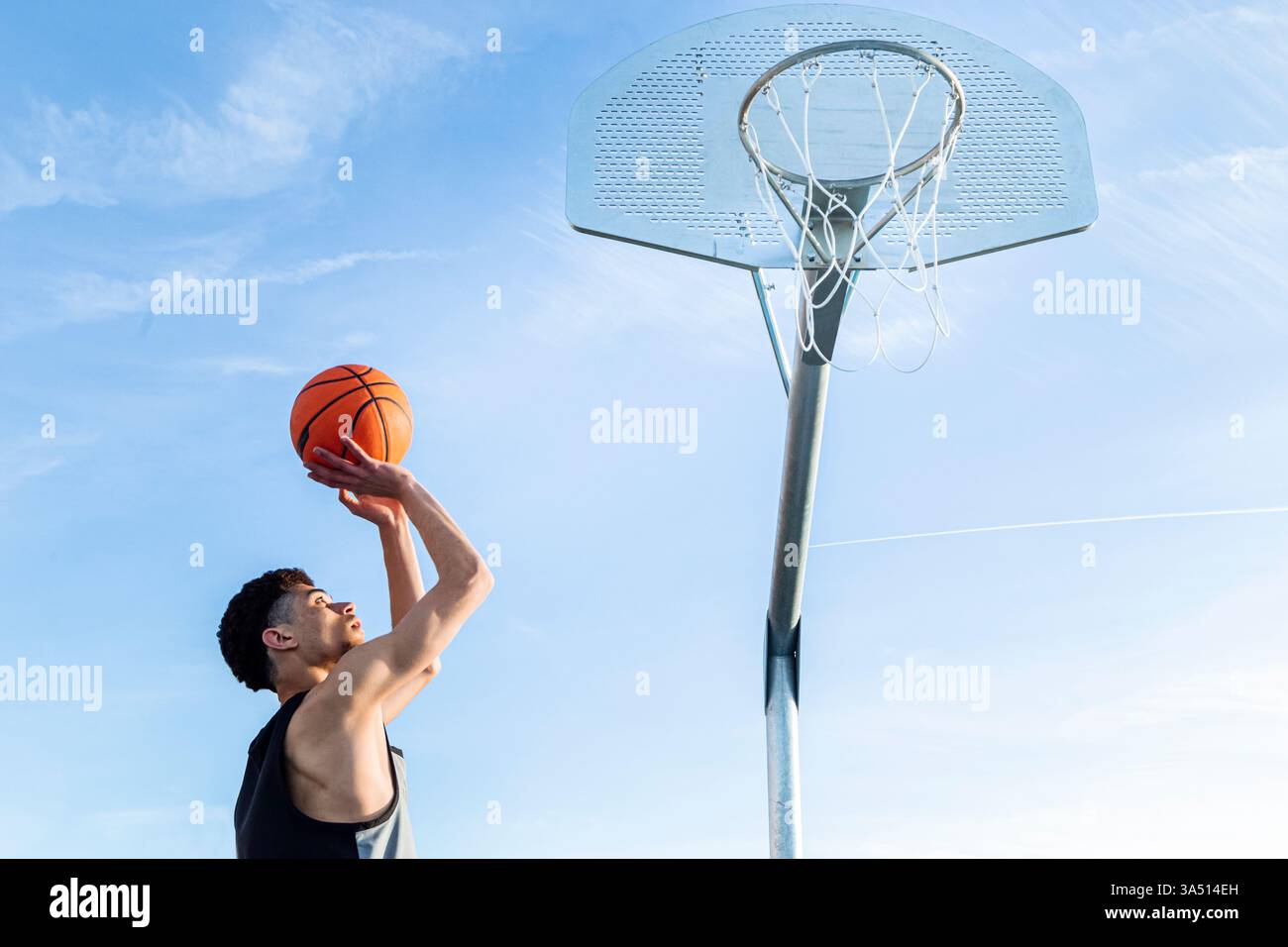 Von unten wirft ein sportlicher Mann an einem sonnigen Sommertag unter blauem Himmel einen Basketball in Richtung Basketball. Diese dynamische Action-Aufnahme eignet sich für Sport-, Fitness- und Outdoor-Lifestyle-Aktionen. Stockfoto