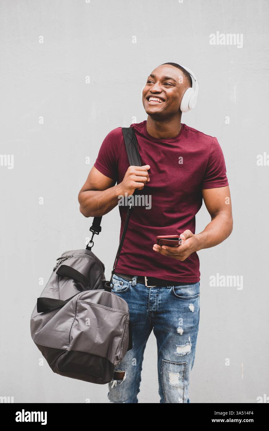 Glücklicher afroamerikanischer Mann, der Kopfhörer trägt, Musik auf seinem Handy hört und eine Tasche trägt. Lässiges Lifestyle-Porträt, geeignet für Technik, Musik und urbane Wohnmotive. Stockfoto
