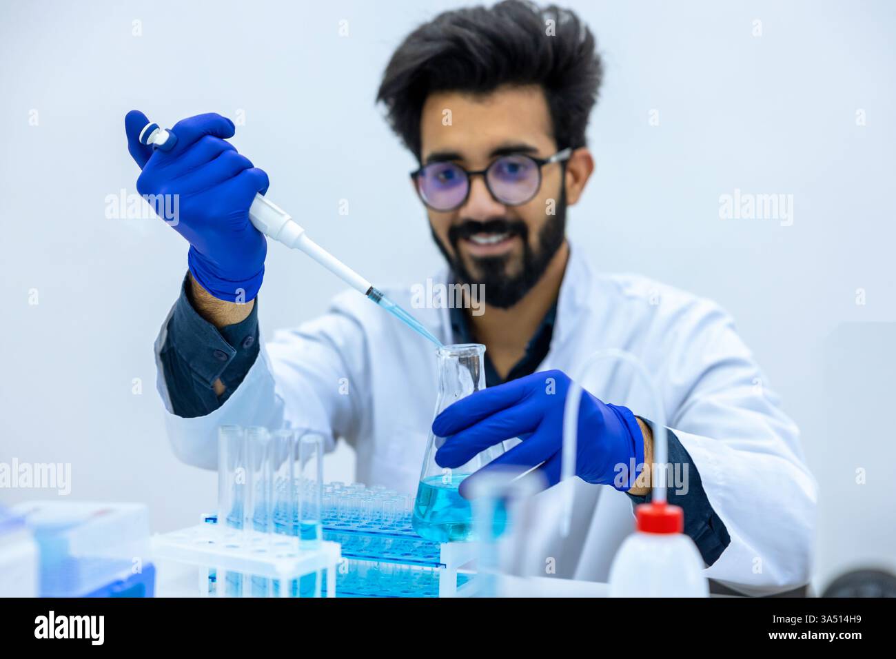 Ein lächelnder indischer Wissenschaftler in einem Labormantel und Handschuhen gießt chemische Flüssigkeit aus einer Pipette in einen Glaskolben an seinem Schreibtisch. Die Szene zeigt Laborarbeiten, Chemie und Biotechnologie, die für wissenschaftliche und pädagogische Funktionen geeignet sind. Es betont präzise Fokussierung und professionelle Arbeit in einer modernen Laborumgebung. Stockfoto