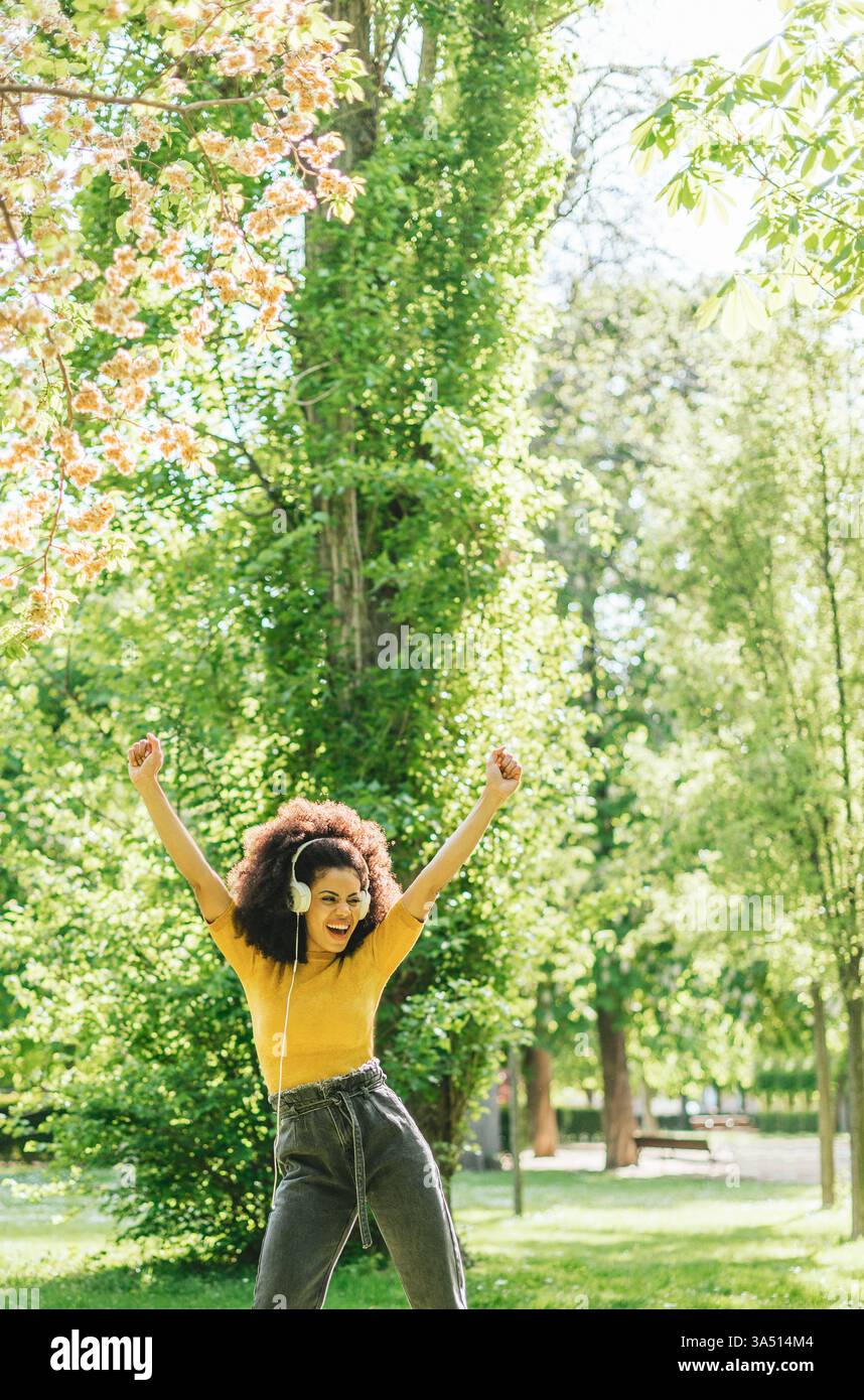 Fröhliche hispanische Frau tanzt in einem Park mit Kopfhörern und erhobenen Armen, während tagsüber Musik die Luft erfüllt. Sie strahlt Energie und Freude aus und hält einen unbeschwerten Moment im Freien fest. Ideal für Lifestyle, Wellness oder Outdoor-Aktivitäten. Stockfoto