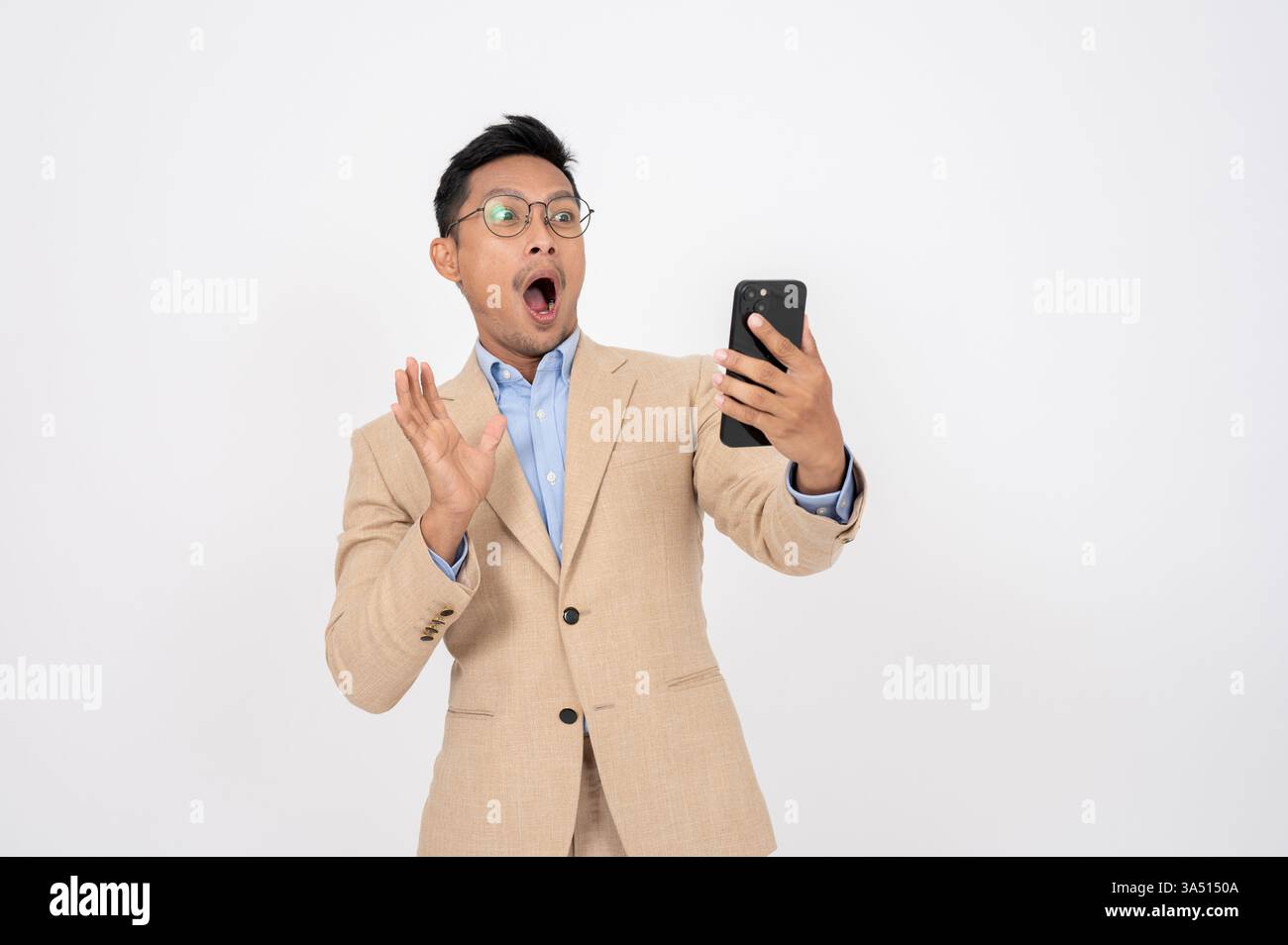 Asiatischer Geschäftsmann auf weißem Atelierhintergrund sieht schockiert und aufgeregt aus, als er auf sein Handy blickt. Dieses Bild mit schneller Reaktion eignet sich hervorragend für Geschäftsankündigungen, Neuigkeiten zu Technologie und Geschäftserfolg. Die Komposition lässt Platz für Marketingnachrichten frei. Stockfoto