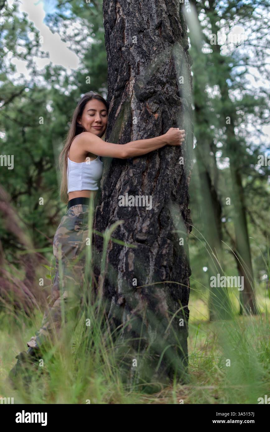 Hispanische Frau, die einen Baumstamm umarmt und die Augen geschlossen hat, während sie im Wald wandert. Ruhiger Naturmoment, der die Verbindung mit der Natur und dem Wohlbefinden betont. Geeignet für Freizeitaktivitäten im Freien, Wanderungen und Freizeitkampagnen in der Natur. Stockfoto