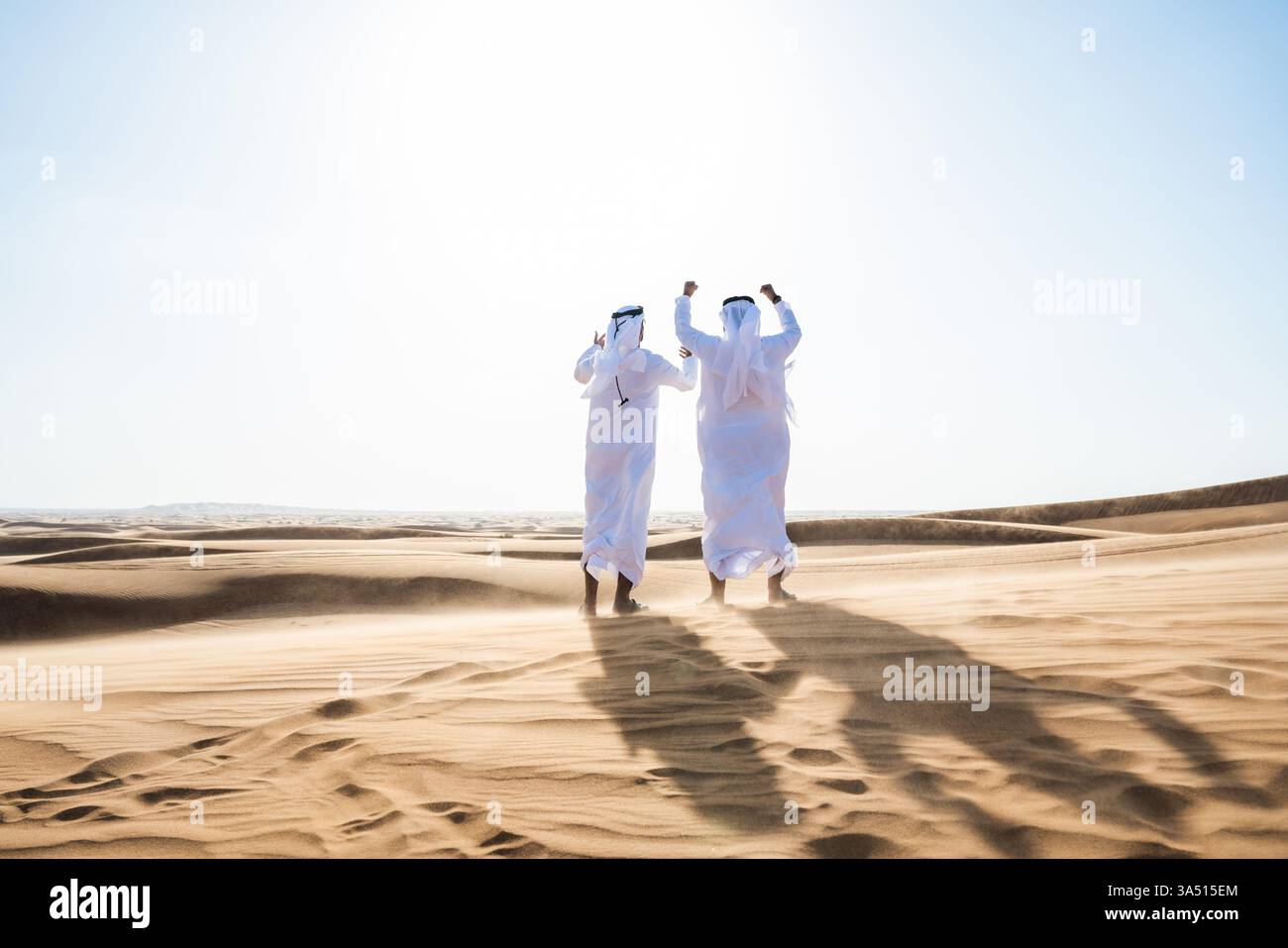 Eine Gruppe von Männern aus dem Nahen Osten und Indien in Kanduras, die sich in einer sonnendurchfluteten Wüste die Hände heben. Ein symbolisches Image für globale Teamarbeit, kulturelle Vielfalt und regionale Geschäftsthemen. Geeignet für Unternehmens-, Reise- und Kulturmarketing. Stockfoto