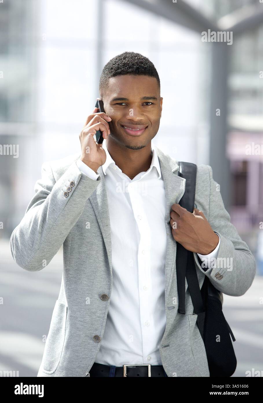 Porträt eines stilvollen jungen Schwarzen Mannes mit einer Tasche, die auf einem Handy spricht. Ideal für Reisen, Pendlerfahrten oder urbane Lifestyle-Kampagnen. Die lässige Jacke und der selbstbewusste Ausdruck des Motivs vermitteln moderne, technisch versierte Professionalität. Stockfoto