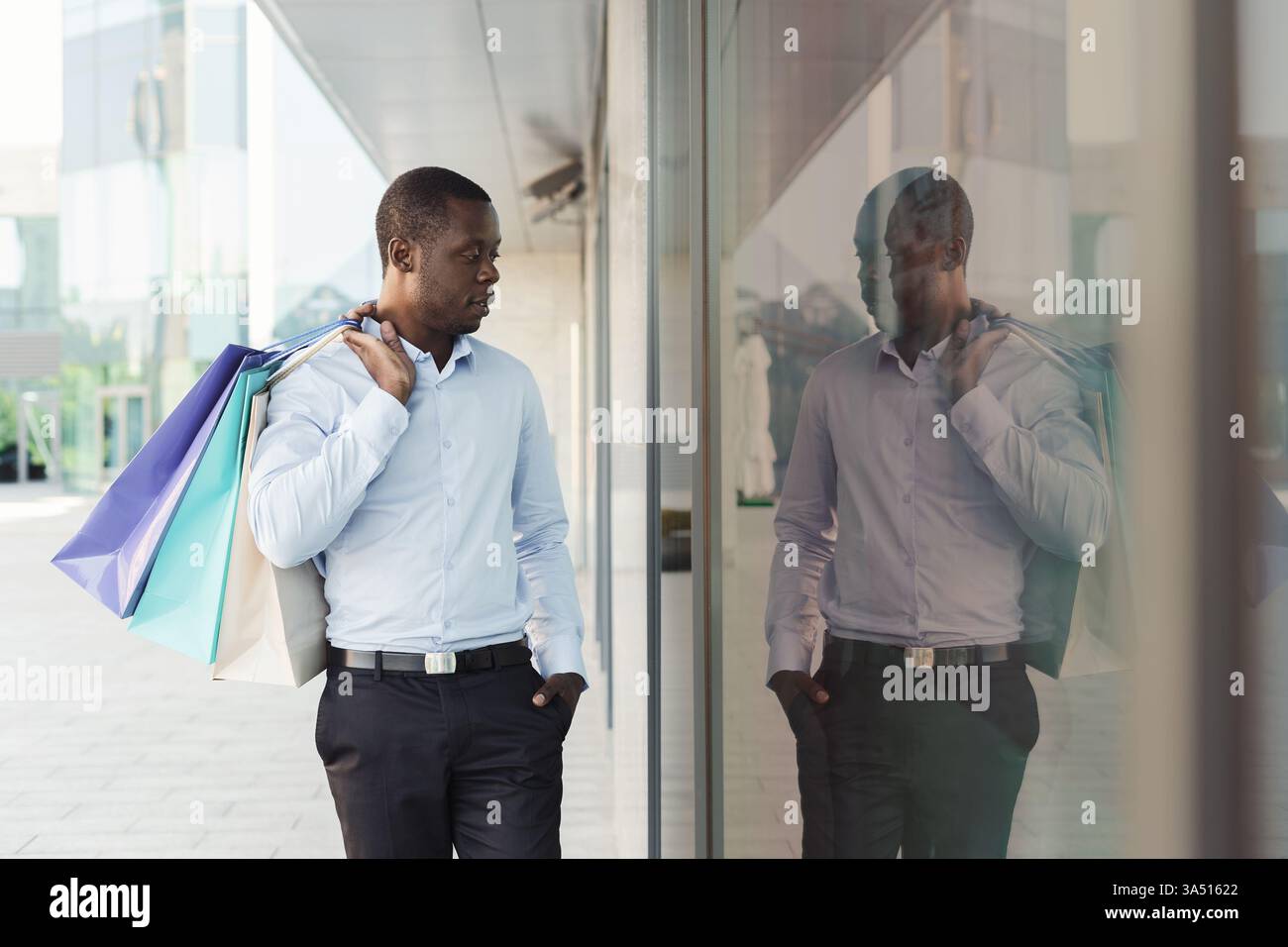 Fröhlicher Black man spaziert entlang einer modernen Boutique-Straße mit Einkaufstaschen. Sein fröhlicher Ausdruck vermittelt Erfolg und Freude am urbanen Shopping. Ideal für Einzelhandelskampagnen, Mode-Lifestyle und Stadtkonsum. Stockfoto