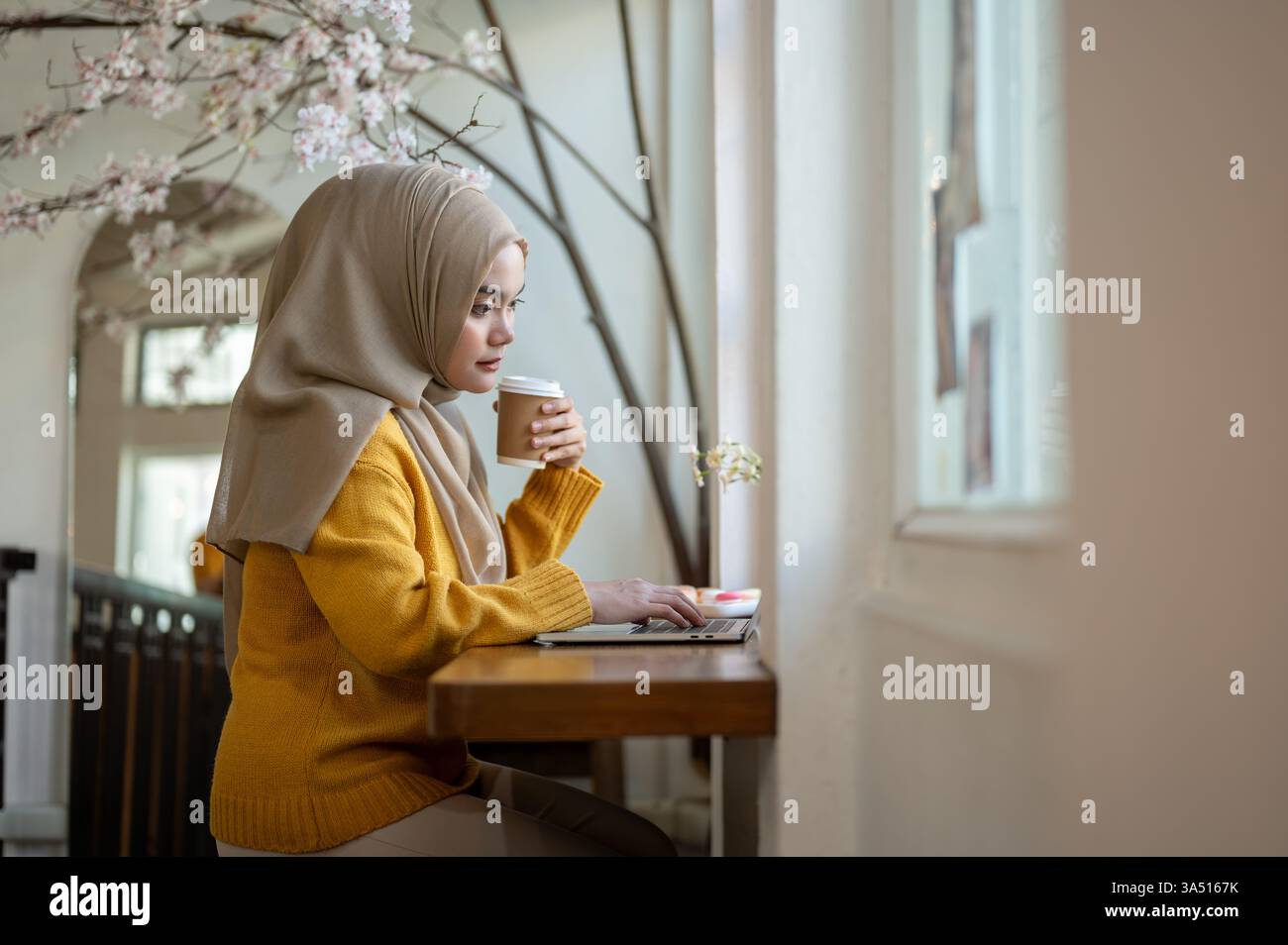 Seitenansicht einer asiatischen muslimischen Frau, die einen Hijab trägt und Kaffee trinkt, während sie einen Laptop in einem Café benutzt. Sie verbindet Arbeit und Freizeit in einem entspannten Café. Ideal für Remote-Arbeiten, freiberufliche Tätigkeiten und multikulturelle Lifestyle-Bilder. Stockfoto