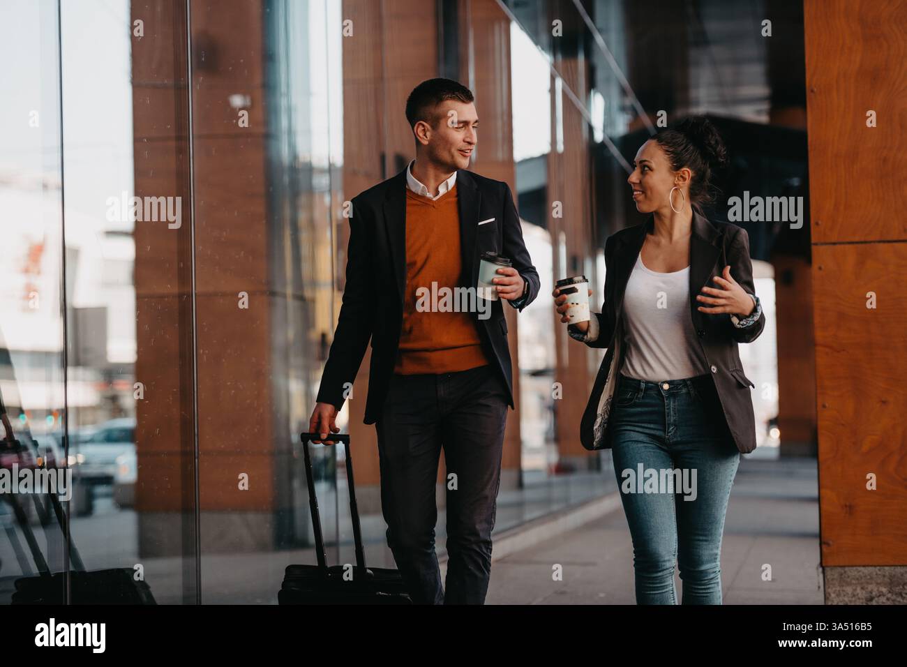 Fröhliche hispanische Geschäftsfrau und kaukasische Geschäftsfrau, die auf Geschäftsreise reist, Kaffee hält und mit Gepäck durch die Stadt spaziert. Ein urbaner, vielseitiger professioneller Moment, der sich für Geschäftsreisen, Tagungen und moderne Kampagnen für den beruflichen Lebensstil eignet. Hervorragend geeignet für Unternehmensservices, Netzwerke und kulturelle Collaboration-Visuals. Stockfoto