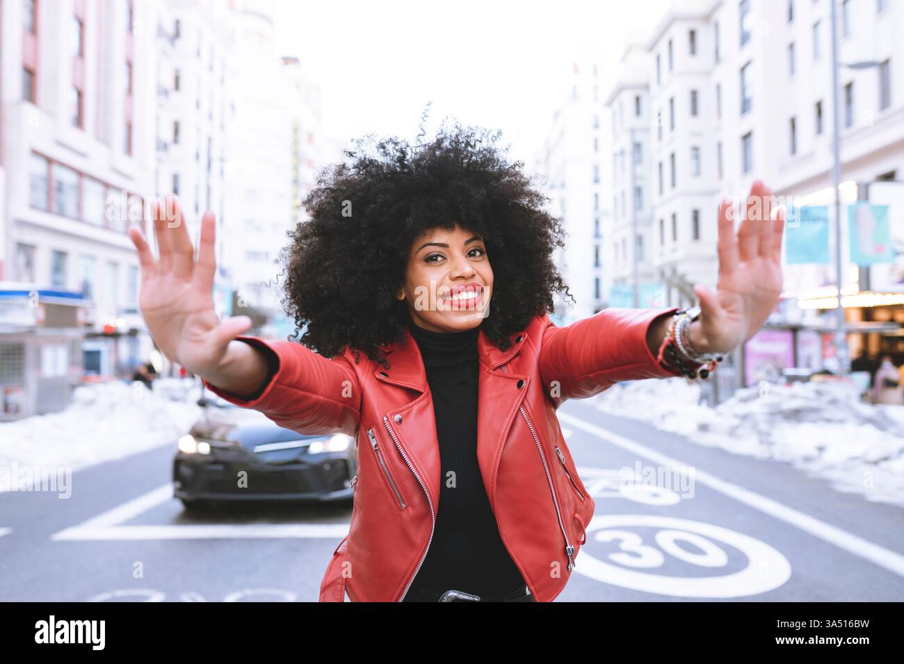 Schwarze Frau auf einer Stadtstraße mit einem Afro steht lächelnd, während sie eine Stop-Geste macht. Dieses ausdrucksstarke städtische Porträt vermittelt Selbstvertrauen und positive Botschaften für Sicherheits-, Bewusstseins- oder Empowerment-Kampagnen. Stockfoto