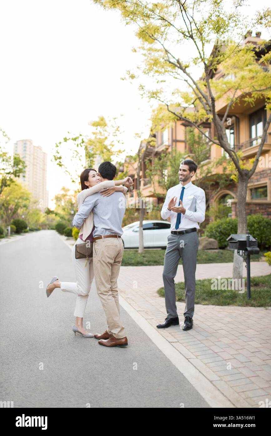 Ein lächelndes chinesisches Paar umarmte sich, nachdem er den Hausschlüssel von einem spanischen Immobilienmakler in einer tagsüber stattfindenden Stadtstraße erhalten hatte. Dieser Moment des Wohneigentums zeigt kulturübergreifende Verbindungen, Zusammenarbeit und Erfolg im Immobilienbereich. Geeignet für Immobilien, Wohnungen und Lifestyle-Geschichten. Stockfoto