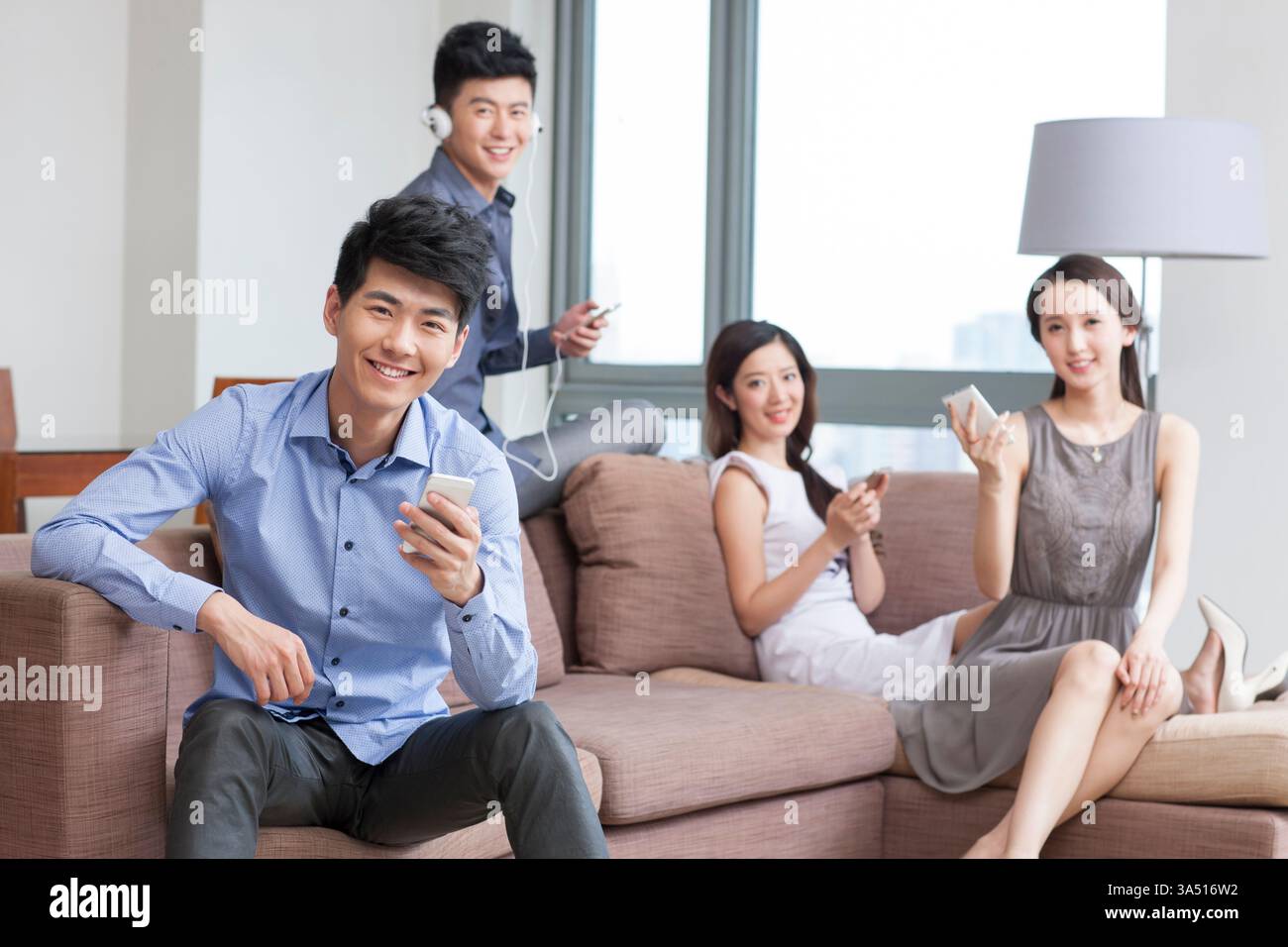 Junge chinesische Freunde, die sich auf einem Wohnzimmer-Sofa entspannen, in ihre Smartphones eingesogen. Die Szene spiegelt modernes Wohnen und digitale Verbindungen wider. Perfekt für soziale Medien, Lifestyle und Technologie. Stockfoto