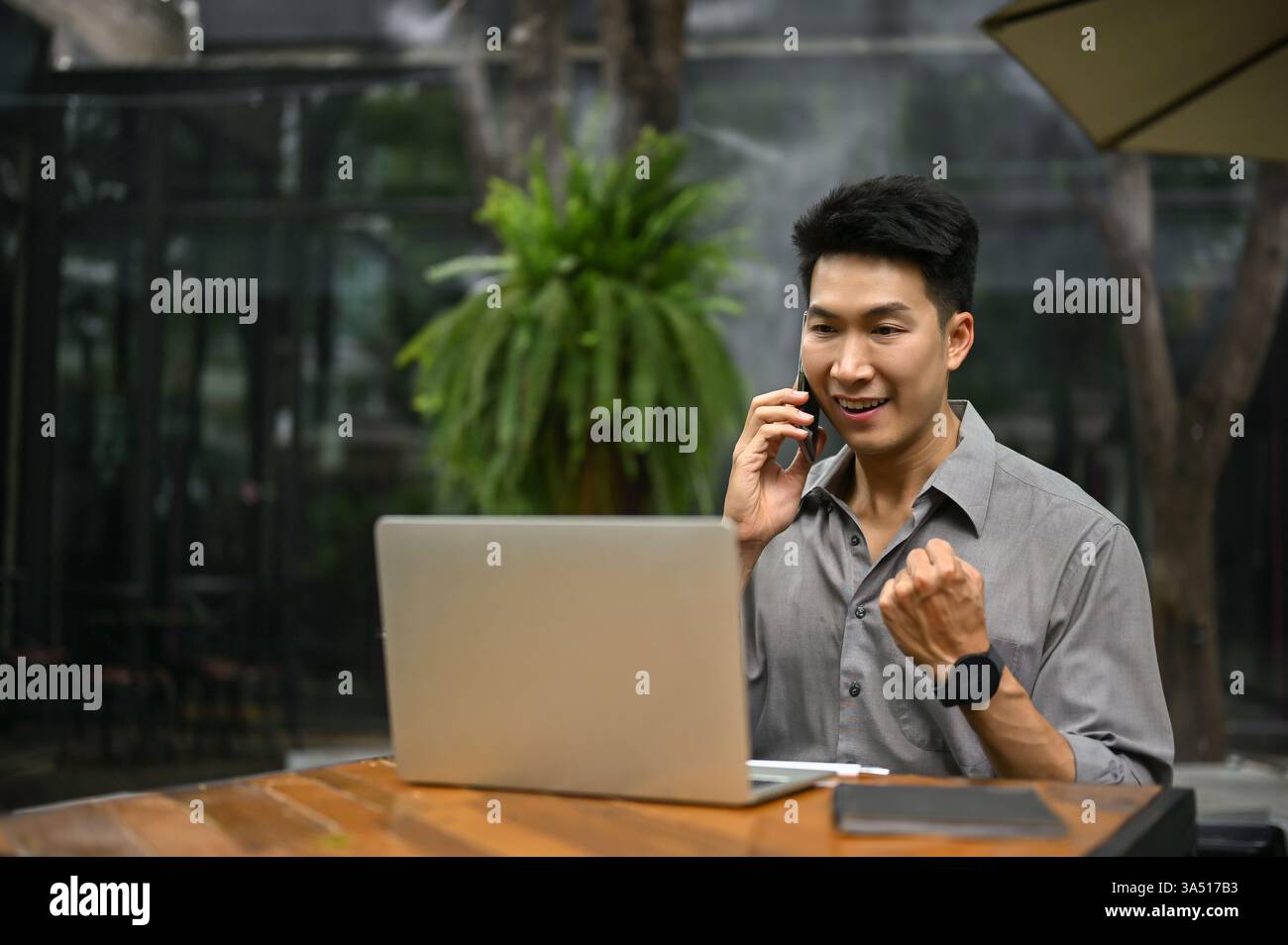 Fröhlicher asiatischer Geschäftsmann der Millennials, der in einem Café gute Nachrichten feiert, während er mit einem Smartphone spricht. Der Moment vermittelt Optimismus und urbane Professionalität. Ideal für Kampagnen für Unternehmen, Start-up- und Remote-Arbeiten. Stockfoto
