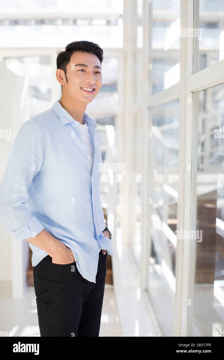 Ein heiterer chinesischer Geschäftsmann steht in einem hellen Büro an einem Fenster mit Händen in der Tasche und blickt weg. Die entspannte Pose vermittelt Selbstvertrauen und eine lässige Geschäftsatmosphäre. Geeignet für Unternehmensprofile, Führungsfunktionen und Arbeitsplatzkultur in modernen Büros. Stockfoto