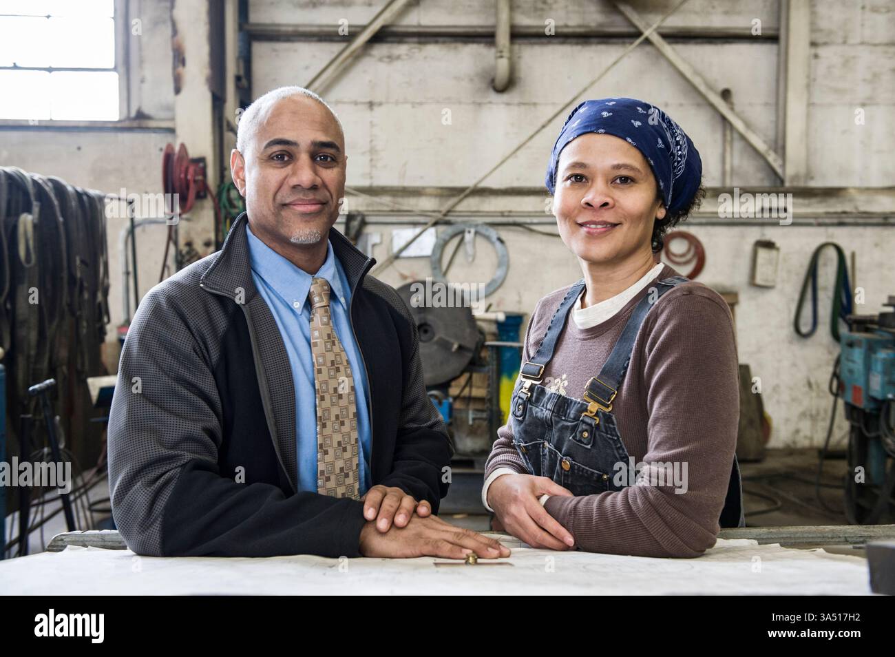 Ein schwarzer Manager und eine asiatische Arbeiterin lehnen sich in industrieller Umgebung an einen Tisch. Die Szene vermittelt Teamarbeit und praktische Zusammenarbeit in einer Fabrik oder Produktionsumgebung. Geeignet für Industrie-, Arbeits- und Teamarbeit am Arbeitsplatz. Stockfoto