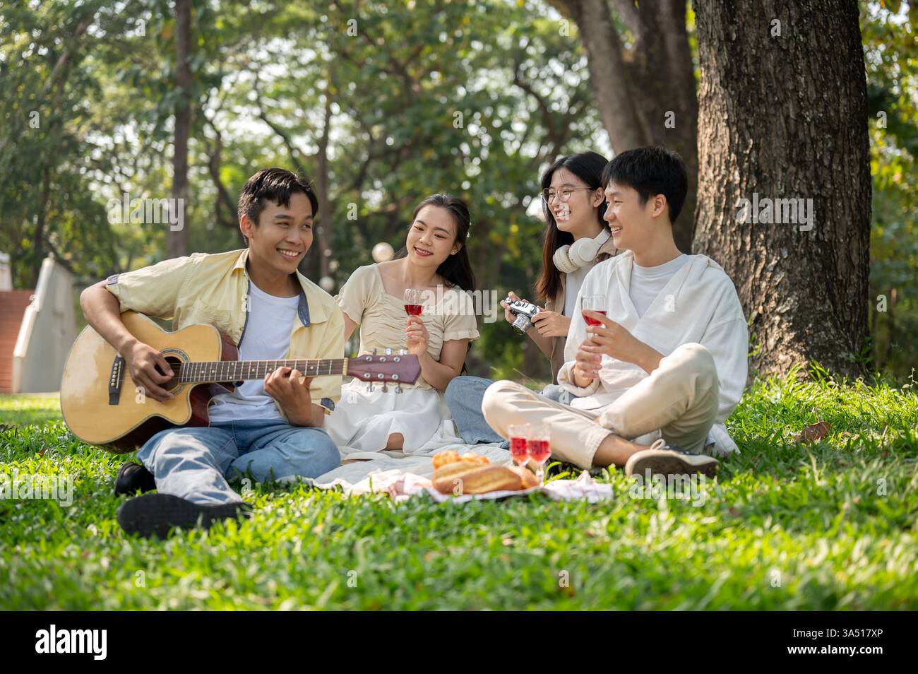 Verschiedene Gruppen asiatischer Freunde picknicken in einem grünen öffentlichen Park mit Gitarrenspiel und Fotos. Eine lebendige Outdoor-Lifestyle-Szene, perfekt für Freundschafts-, Freizeit- und Sommerkampagnen. Stockfoto