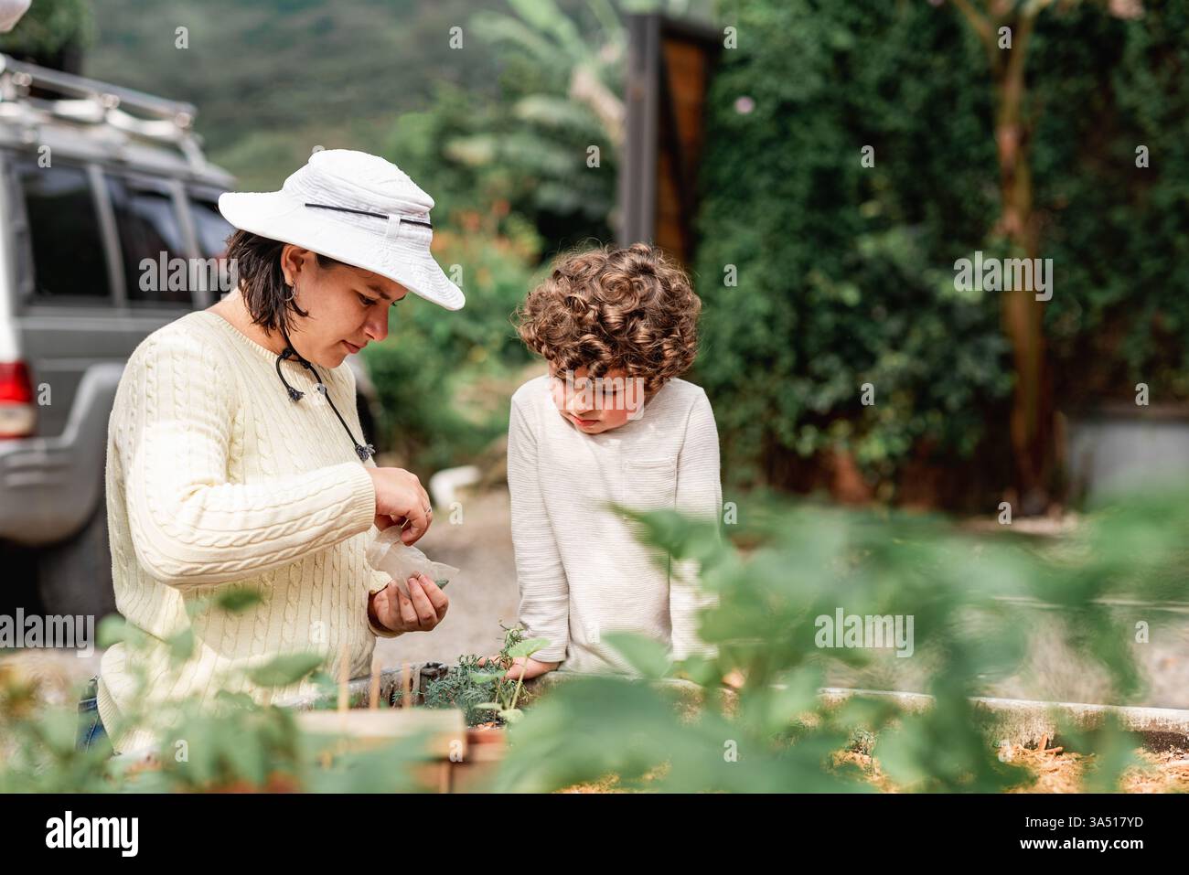 Hispanische Gärtnerin nimmt Samen aus einem Plastikbeutel, um sie mit ihrem kleinen Sohn draußen in ein Gartenbeet zu Pflanzen. Ideal für Familien-, Garten- und Landleben. Authentische, warme Szene, die für Marken geeignet ist, die Lernen im Freien und nachhaltiges Leben feiern. Stockfoto