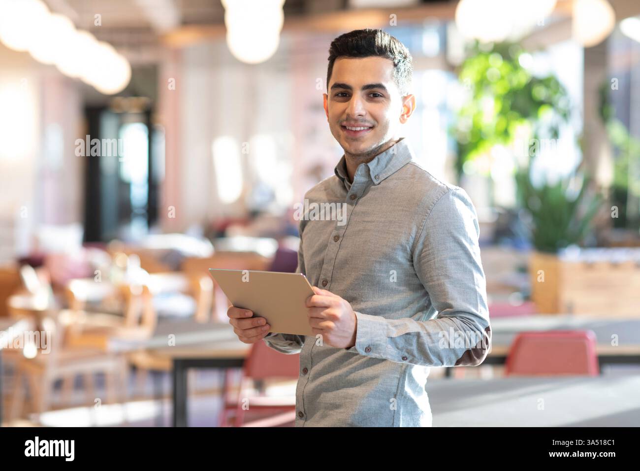Ein lächelnder Mann aus dem Nahen Osten steht in einem Café mit einem Tablet. Das Bild vermittelt digitale Konnektivität und moderne Alltagsmomente. Ideal für Technik, Studentenleben und Gastfreundschaft. Stockfoto