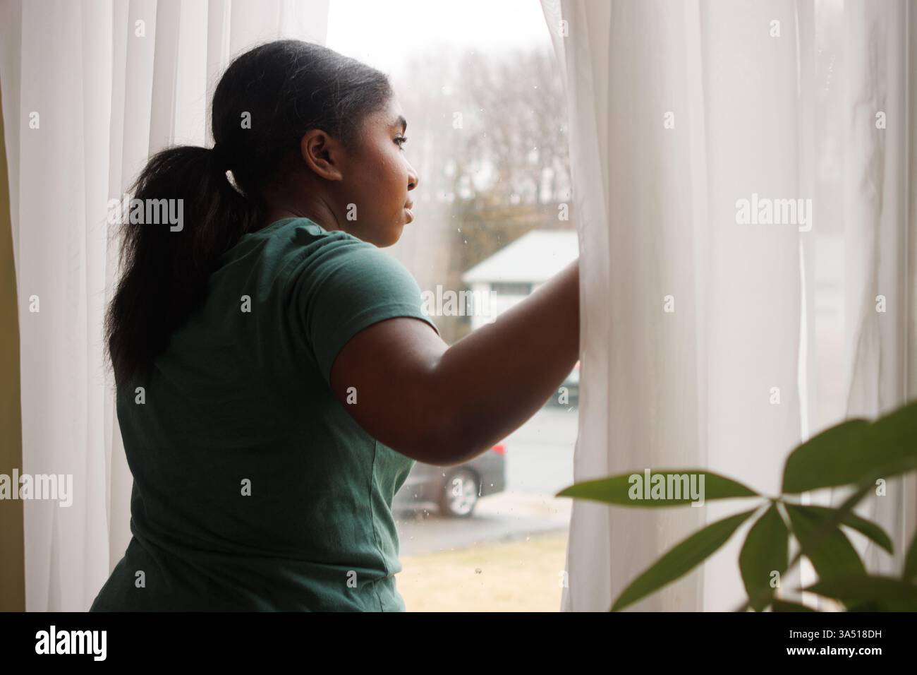 Schwarze Teenagerin steht zu Hause an einem Fenster und blickt mit hoffnungsvollem Ausdruck nach draußen. Ideal für Jugendliche, das Heim- und zukünftige Sehnsüchte. Stockfoto