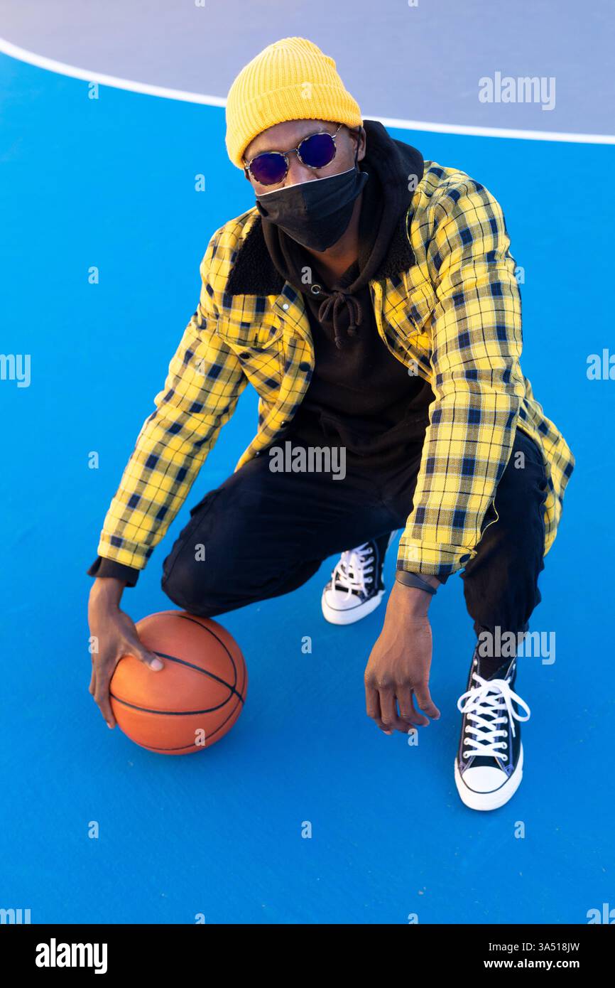 Schwarzer männlicher Basketballspieler mit Maske auf dem Platz. Dieses sportliche Image aus der Pandemie betont sportlichen Fokus und Sicherheit und eignet sich für Fitnesskampagnen, Teamaktionen oder sportliche redaktionelle Zwecke. Ein kühnes Porträt von Entschlossenheit und Sport. Stockfoto