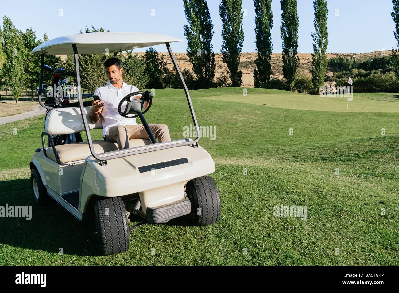 Hispanischer Golfspieler mit Smartphone, während er an einem sonnigen Tag mit einem Golfwagen über einen grünen Golfplatz fährt. Die Szene verbindet Sport, Technologie und Freizeit in einer Outdoor-Umgebung. Geeignet für Golf-Lifestyle, mobile Technologie und Outdoor-Branding. Stockfoto