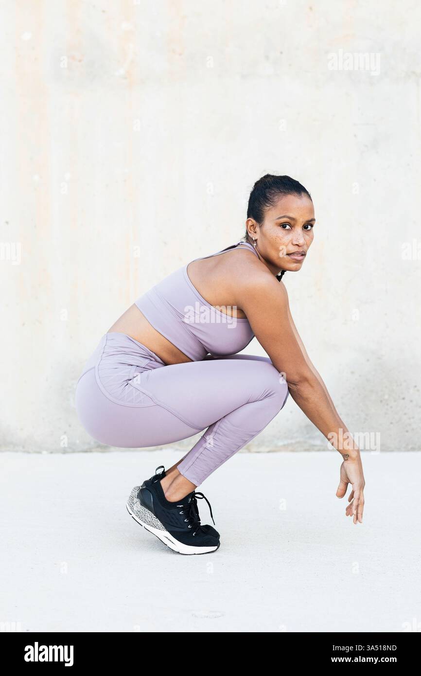 Afroamerikanerin in Sportkleidung hockt draußen auf Zehenspitzen gegen eine Wand. Fitness- und Activewear-Bilder, die Bewegung und Wellness hervorheben. Ideal für gesundes Leben, Sport und Trainingskampagnen. Stockfoto