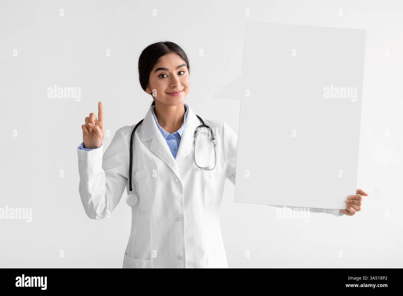 Lächelnde indische Ärztin in Uniform hält ein Banner mit Kopierraum hoch, ideal für Gesundheitskampagnen und COVID-19-Bewusstsein. Der saubere Studiohintergrund betont Professionalität und klare Botschaften. Stockfoto