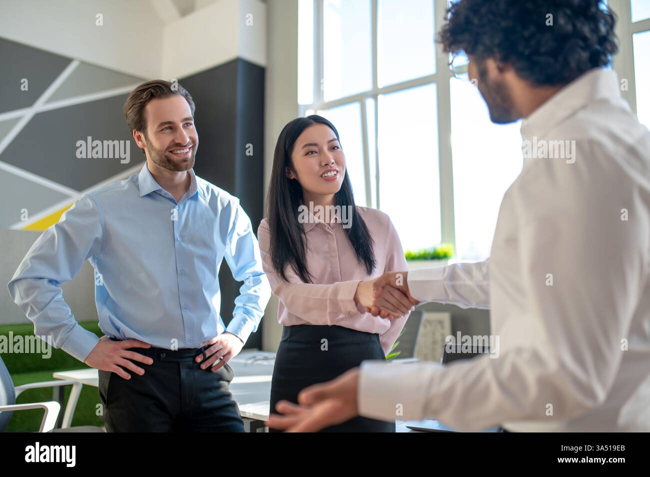 Lächelnde asiatische Geschäftsfrau schüttelt einem indischen Geschäftsmann die Hand, während ein kaukasischer Kollege in einem hellen Büro zusieht. Die internationale Zusammenarbeit vermittelt Professionalität und positive Geschäftsbeziehungen. Stockfoto