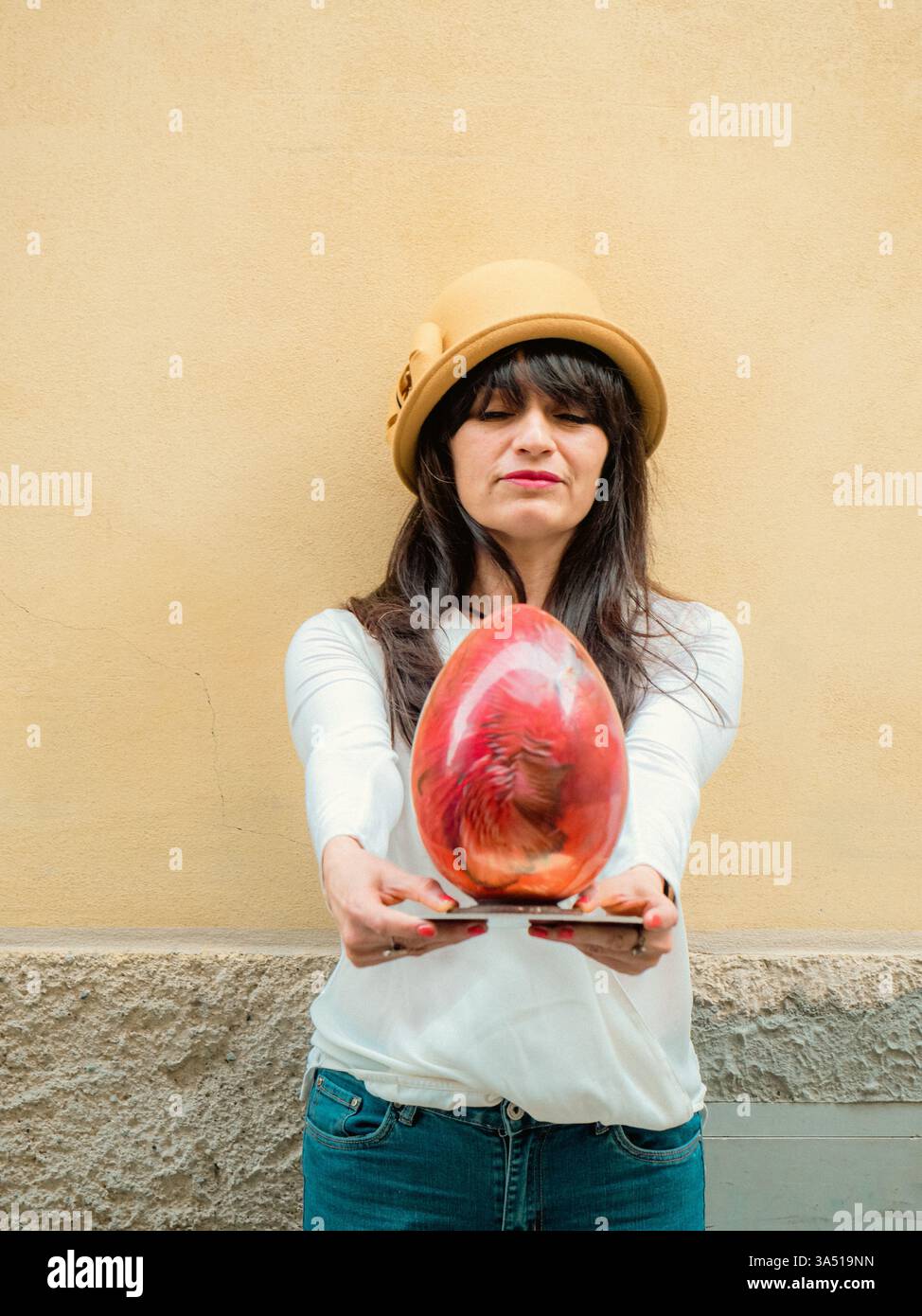 Eine hispanische Erwachsene Frau steht draußen und hält ein rotes, handwerkliches Osterei aus italienischer Schokolade in der Hand. Eine festliche Osterszene, die für saisonale Feierlichkeiten und Feiertagsdekor geeignet ist, mit Kopierraum für Schlagzeilen. Perfekt für Geschenkideen zu Ostern, Frühlingskampagnen und Marketing zum Thema Feier. Stockfoto