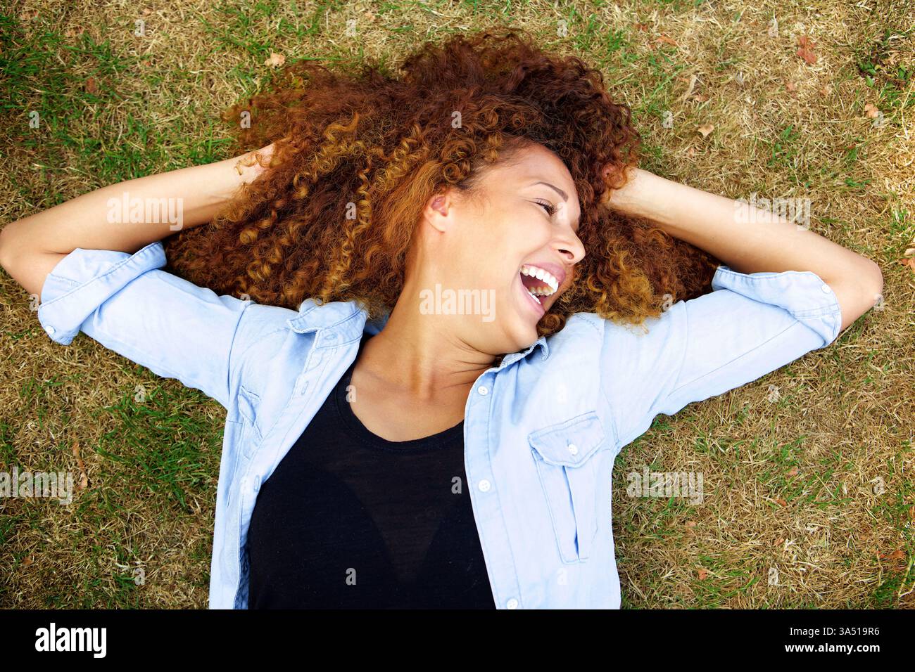 Porträt einer jungen Frau, die im Freien auf Gras lacht. Die sorglose, fröhliche Pose macht ihn ideal für Wellness, Natur und positive Lifestyle-Themen. Ideal für Redakteure, die ein brillantes, aufmunterndes Bild von Selbstvertrauen und Glück benötigen. Stockfoto