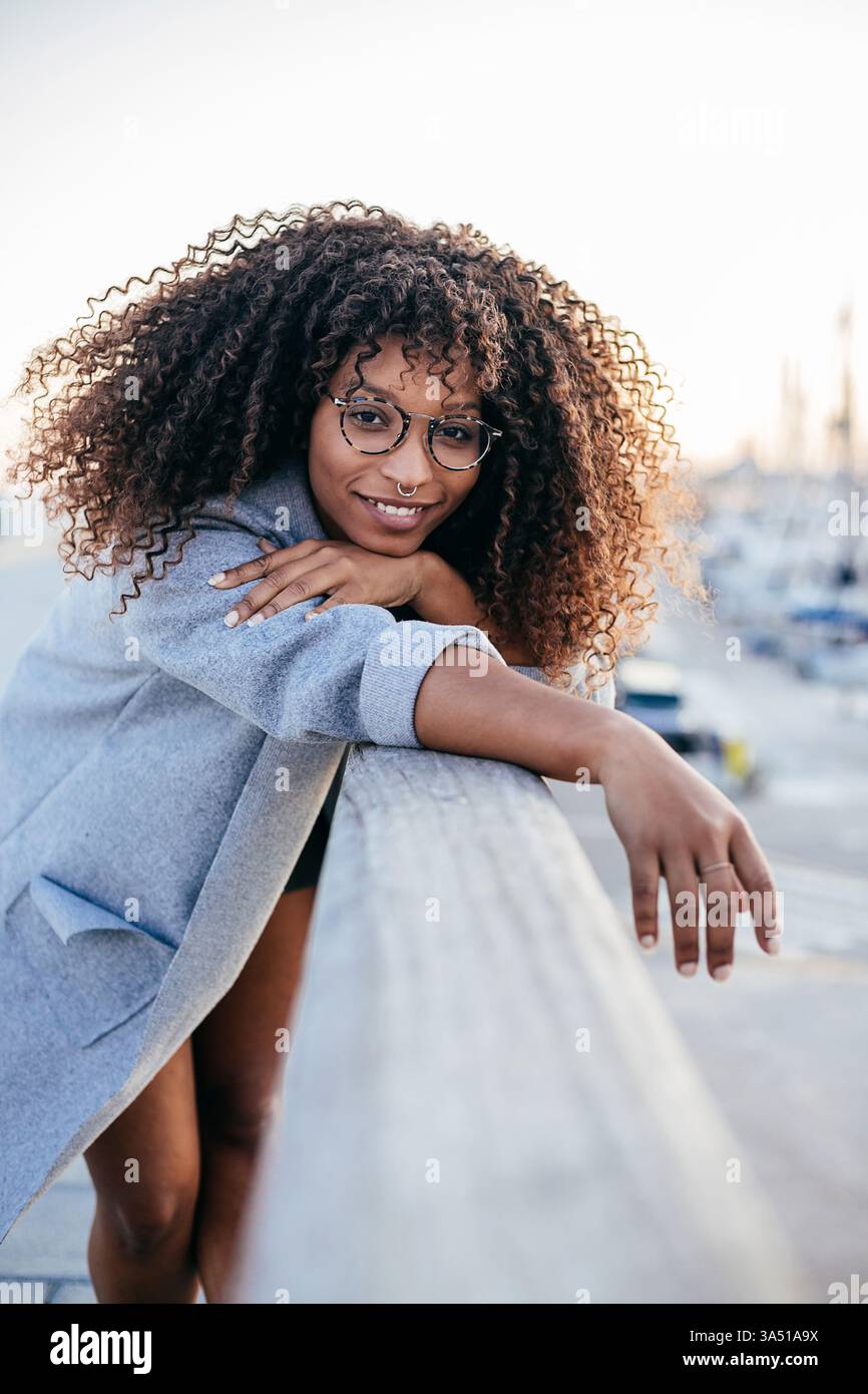 Lächelnde Afroamerikanerin mit lockigen Haaren lehnt sich auf einen Zaun neben dem Hafen. Dieses Porträt im Freien fängt einen fröhlichen, entspannten Moment in einer Umgebung am Wasser mit modernem urbanem Flair ein. Perfekt für Lifestyle-Accessoires, die glückliche, selbstbewusste Frauen im legeren Sommer-Style feiern. Stockfoto