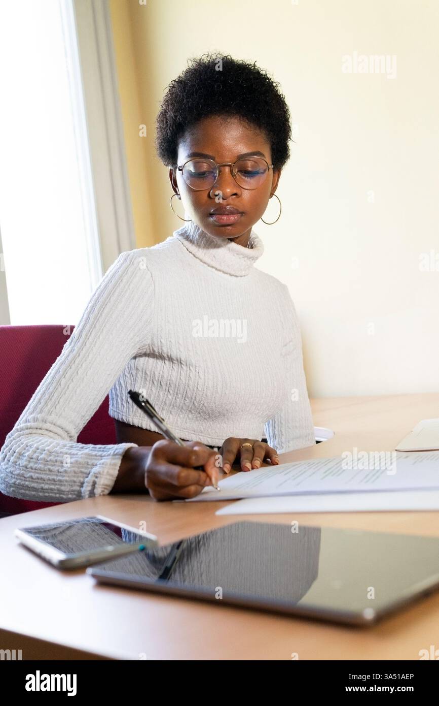 Schwarze Frau signiert Dokumente an einem Tisch mit Tablet und Smartphone in einem modernen Büro. Das professionelle Umfeld kommuniziert die Unternehmensverwaltung und den digitalen Workflow. Geeignet für Unternehmens-, Karriere- und Frauenthemen. Stockfoto