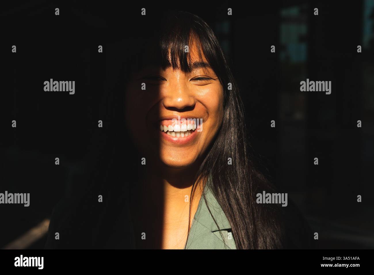 Fröhliche asiatische Frau im Schatten mit Sonnenstrahlen im Gesicht. Sie ist im Freien bei natürlichem Licht und schafft ein helles, positives Porträt. Gut für Lifestyle- und Outdoor-Kampagnen. Stockfoto