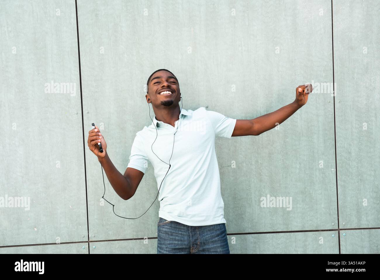 Porträt eines fröhlichen jungen afroamerikanischen Mannes, der Musik mit Ohrstöpseln genießt, während er auf sein Mobiltelefon schaut. Ein lässiges Lifestyle-Image, das perfekt für Musik, Technologie und Smartphone-Kampagnen geeignet ist. Stockfoto