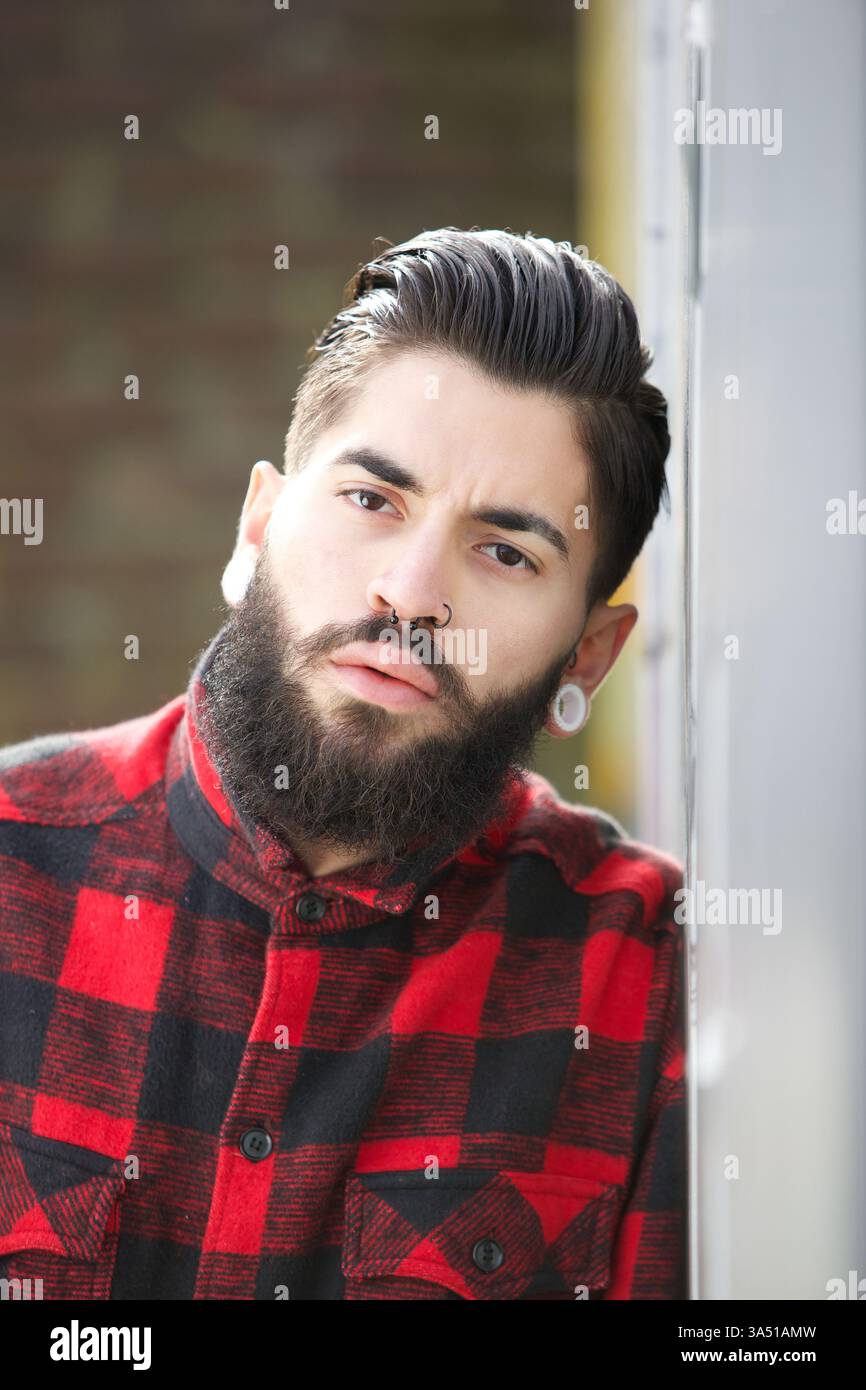 Nahporträt eines bärtigen Mannes mit Piercings, das einen auffälligen, modernen Look präsentiert. Das städtische Feeling und die stilvollen Details machen es für Mode- und Porträtfotografie geeignet. Ideal für Mode, Lifestyle und trendige urbane Themen. Stockfoto