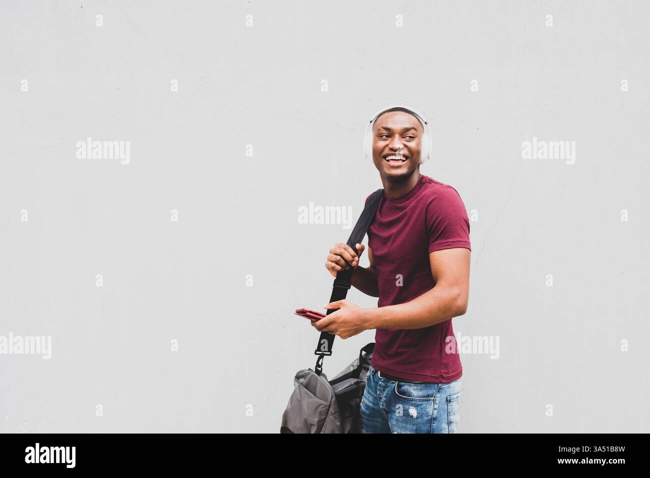 Lässiges Porträt eines lächelnden afroamerikanischen Mannes, der mit einer Tasche geht, Kopfhörer trägt und ein Smartphone benutzt. Seine entspannte Lifestyle-Atmosphäre macht es für Apps, Musik und Technologiekampagnen geeignet. Natürliche Komposition mit Platz für Text. Stockfoto