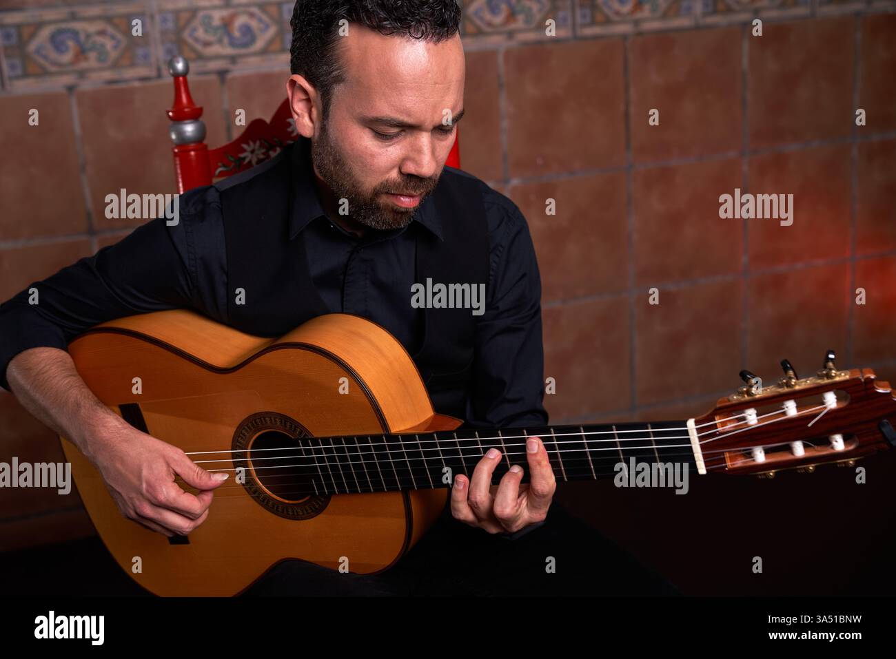 Der talentierte bärtige männliche Musiker in einem schwarzen Rollkragen spielt eine Akustikgitarre, sitzt in der Nähe einer gekachelten Wand. In diesem Innenbereich wird ein konzentrierter Moment des Trainings und der Kreativität festgehalten. Perfekt für Musik, Lifestyle und künstlerisches Branding mit einer warmen, intimen Stimmung. Stockfoto