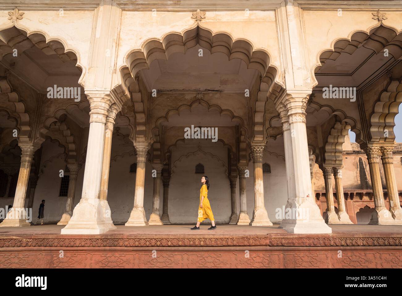 Asiatische Touristin in einem leuchtend gelben Kleid, die an alten Säulen in einer bogenförmigen Passage eines historischen Gebäudes vorbeigeht. Ideal für Reise- und Kulturfotografie, Architekturmerkmale und Reiseführer. Dieses Bild eignet sich für kulturelle Sehenswürdigkeiten und Erkundungsthemen. Stockfoto
