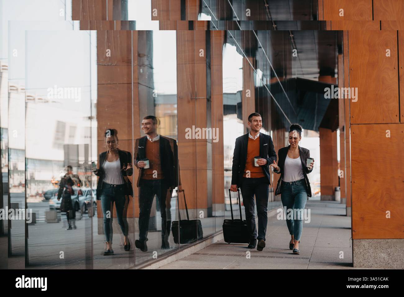 Fröhliche hispanische Geschäftsfrau und kaukasischer Geschäftsmann reisen auf Geschäftsreise, schlendern mit Kaffee in der Hand und Gepäck. Die städtische Umgebung erinnert an Geschäftsreisen, Meetings und professionelle Planung. Stockfoto