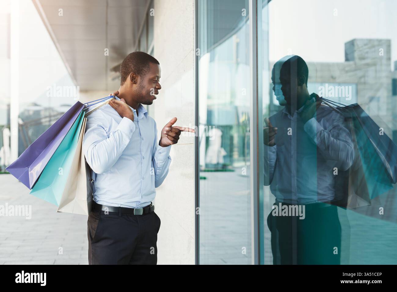 Glücklicher afroamerikanischer Mann, der mit Einkaufstaschen entlang einer modernen Stadtstraße spaziert. Ein Stadtbild mit Einzelhandels- und Modemerkmalen zeigt Erfolg und Stil. Perfekt für Kampagnen zu Shopping, Unternehmergeist und urbanem Lebensstil. Stockfoto