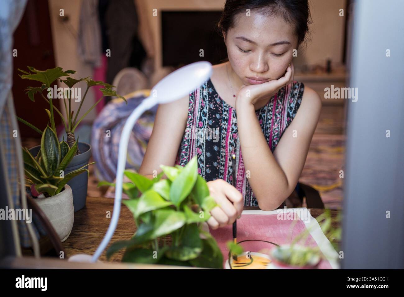 Asiatische Künstlerin sitzt an einem Holztisch und malt mit einem Pinsel auf Papier. Kreative Studioszene für Kunst-, Bastel- und Designthemen. Ideal für kleine Unternehmen und handgefertigte Produktinhalte. Stockfoto
