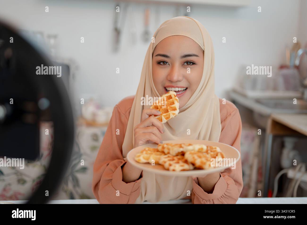 Die asiatische Muslimin, eine Lifestyle-Influencerin und Food-Bloggerin, nimmt ein Video auf, während sie in einer Küche Waffeln isst. Sie teilt Inhalte über Kochen, Frühstück und Social-Media-Streaming mit einem Smartphone und einer Kamera. Ihre Inhalte umfassen Kochen, Rezeptaustausch und Live-Streaming für Follower. Stockfoto