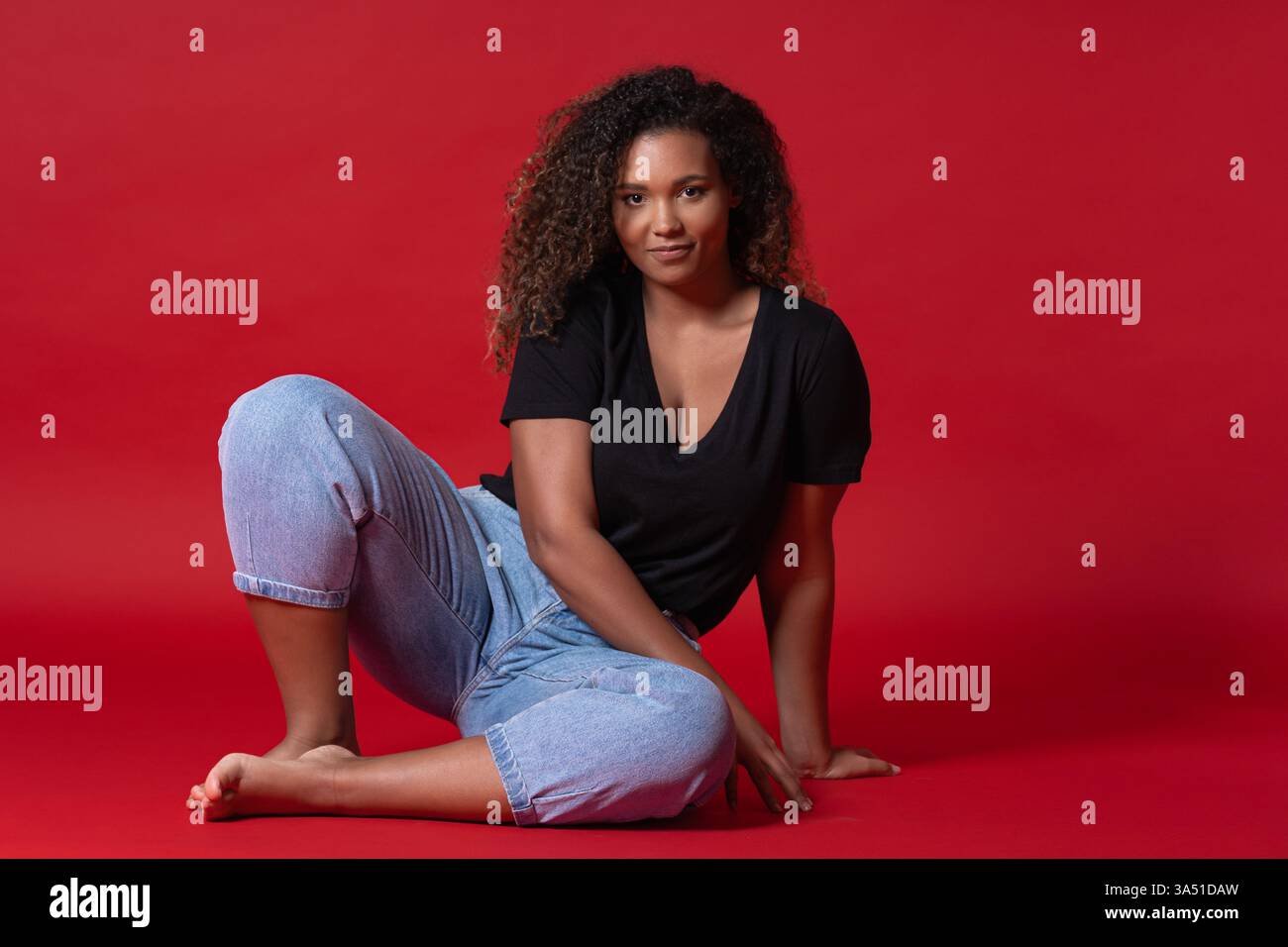 Lässiges Porträt einer stylischen Afroamerikanerin in schwarzem Hemd und Jeans, barfuß vor einem auffälligen roten Studiohintergrund. Sie strahlt Selbstvertrauen und Wärme aus und reflektiert körperpositive Mode und Alltagsstyle. Ideal für Lifestyle- und Modekampagnen mit bequemer Freizeitkleidung. Stockfoto