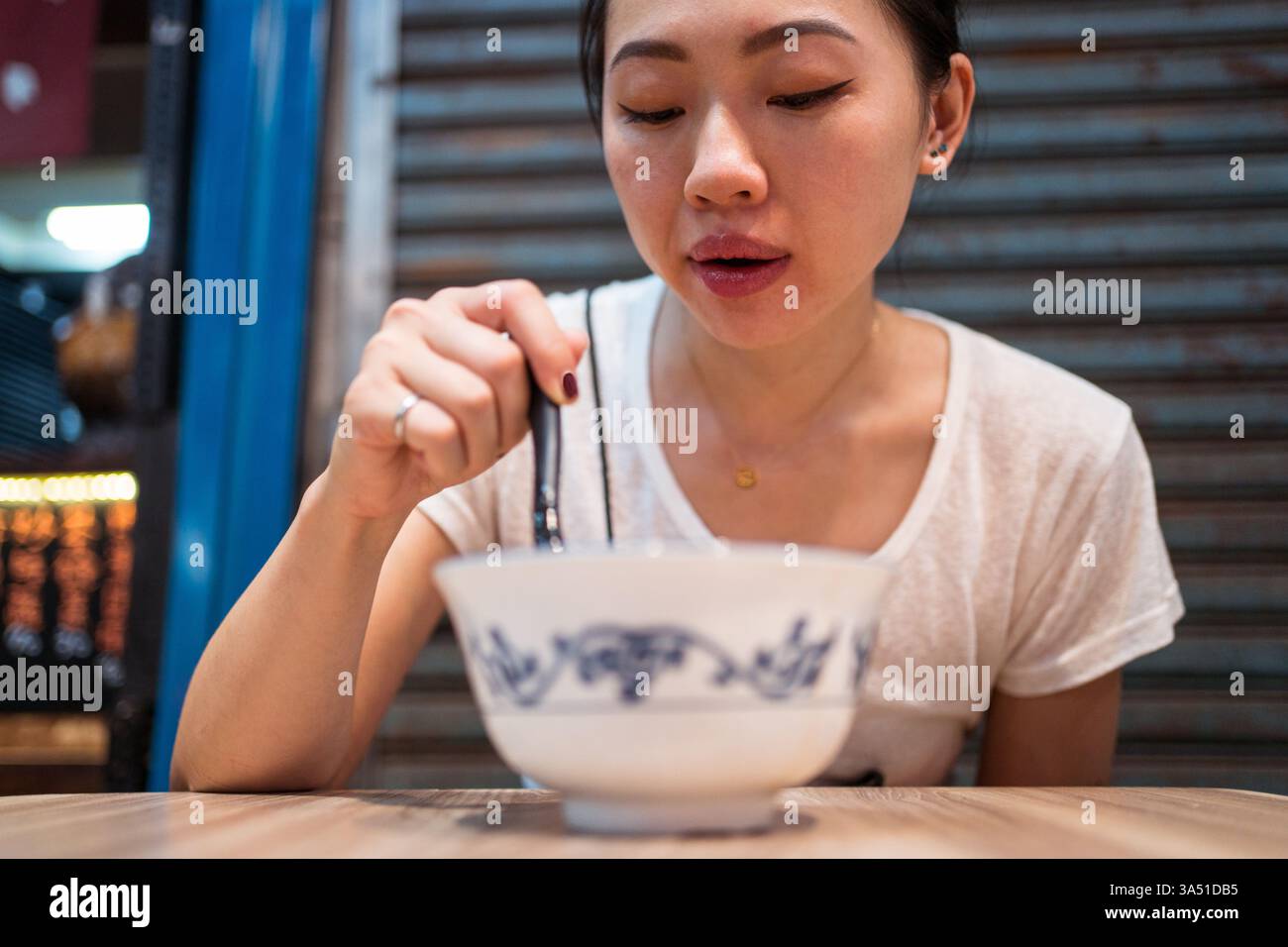 Die junge asiatische Frau sitzt an einem Holztisch und genießt taiwanesische Nudelsuppe. Das zwanglose Dining-Moment bietet authentische asiatische Küche mit einer geschmackvollen Schüssel. Geeignet für Speisen, Cafékultur und asiatische Küche Angebote. Stockfoto