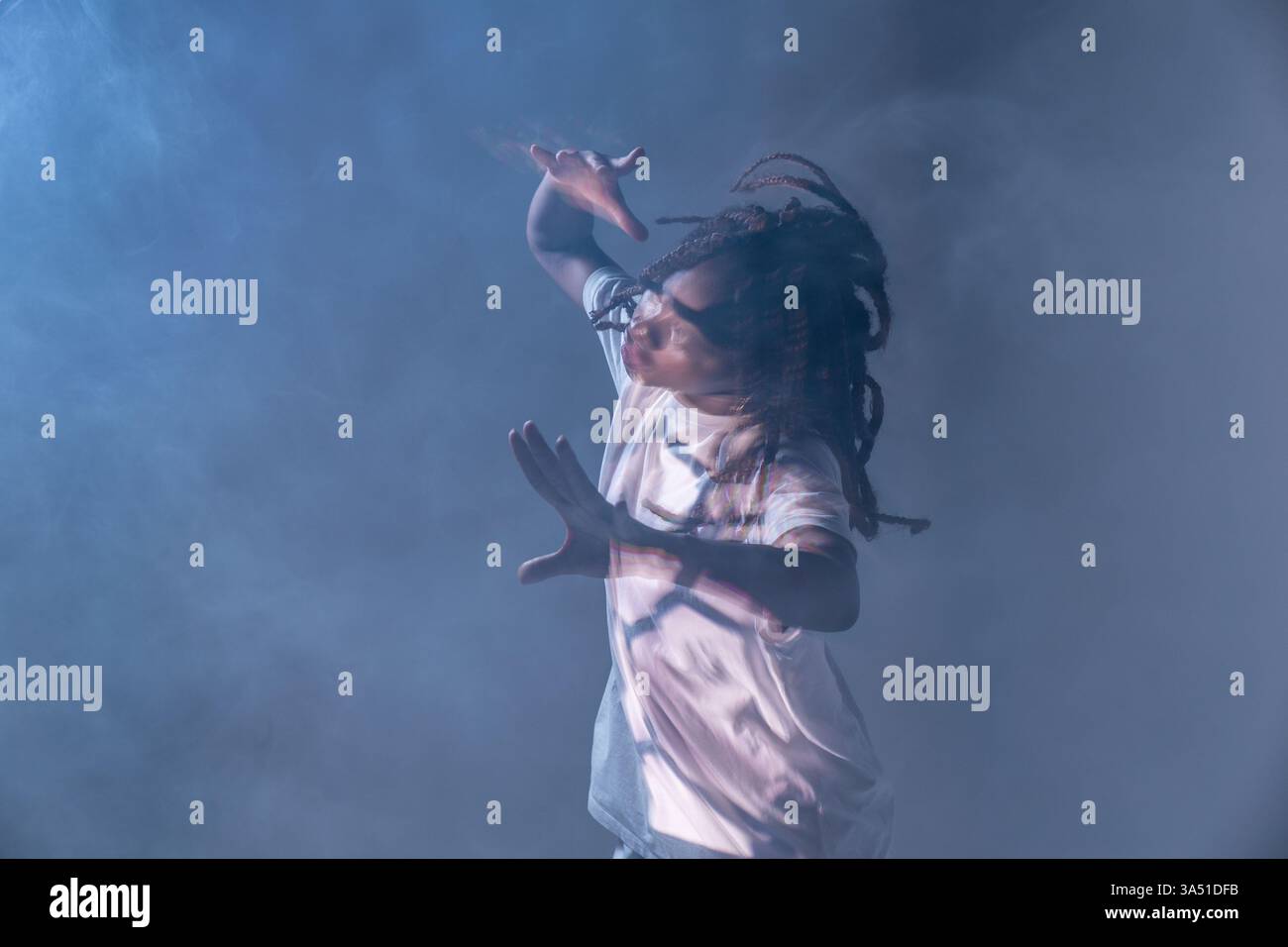 Dramatisches Bild einer afroamerikanischen Tänzerin mit Dreadlocks, die vor blauem Hintergrund mit Rauch tanzen. Die dynamische Pose und die lebendigen Farben vermitteln Energie und Jugendlichkeit. Stockfoto