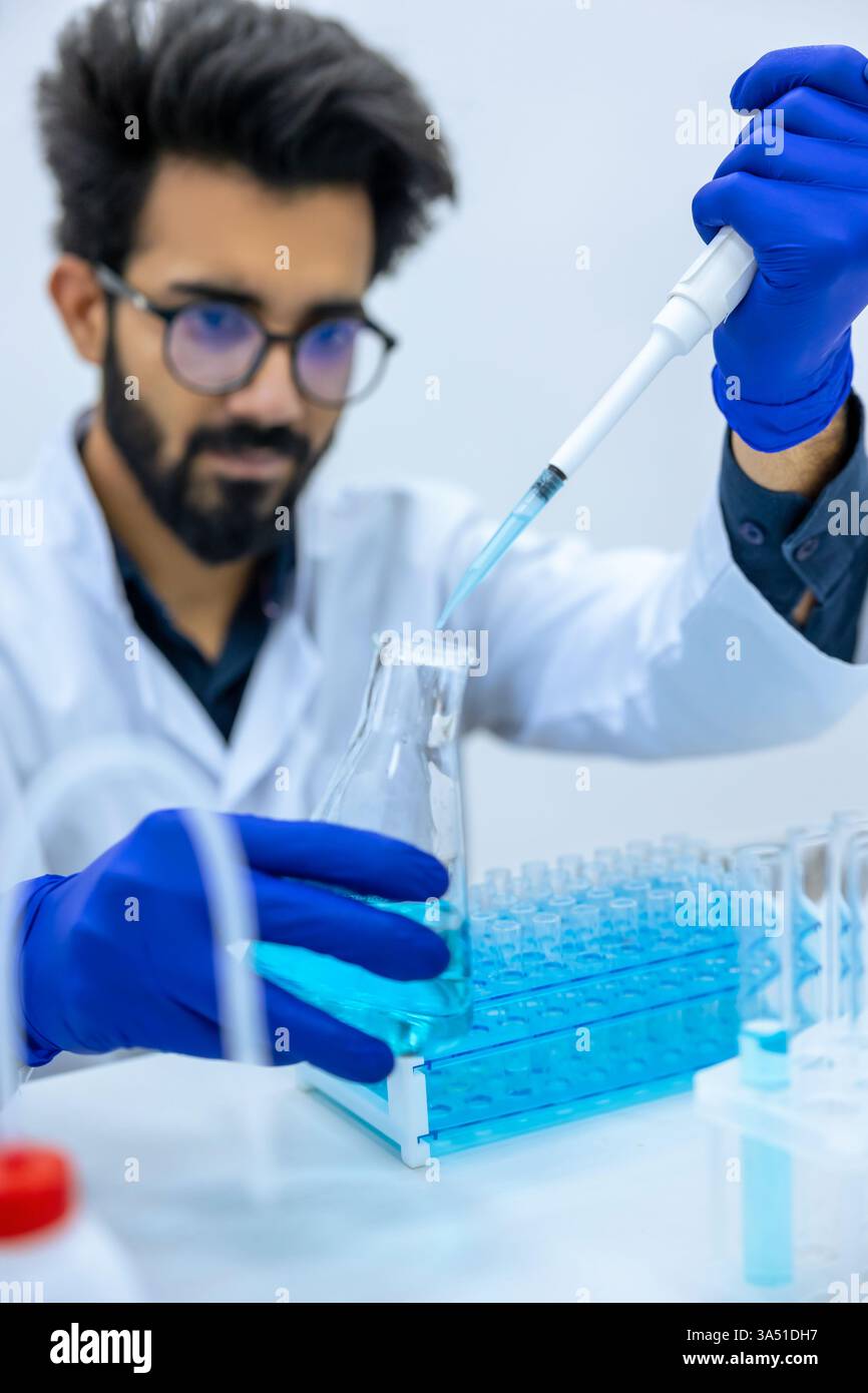 Konzentrierter indischer Wissenschaftler mit Labormantel und Latexhandschuhen, der chemische Flüssigkeit aus der Pipette in den Glaskolben gießt, während er ... an seinem Schreibtisch saß Stockfoto