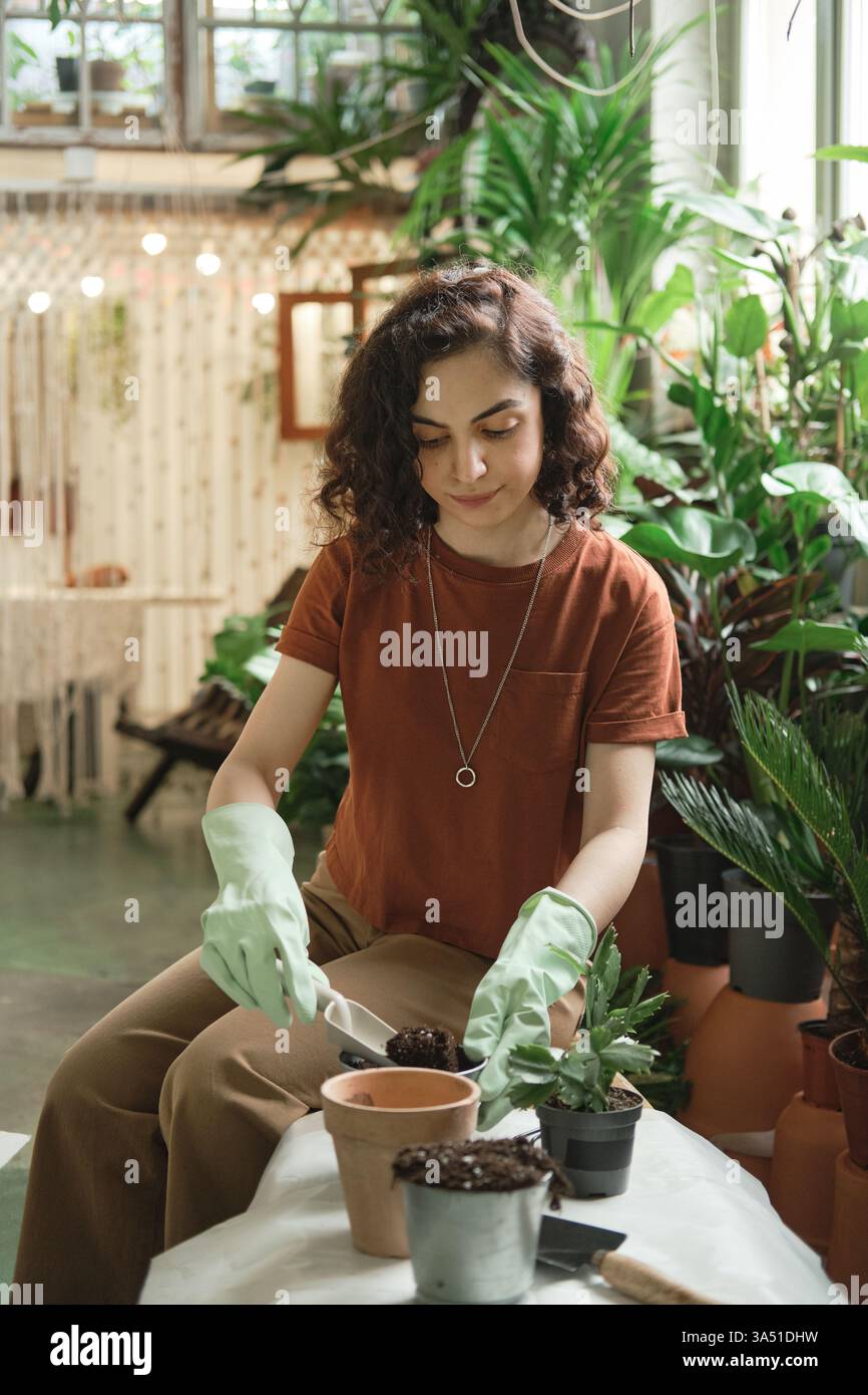 Eine junge Frau in lässiger Kleidung pflanzt eine grüne Pflanze in einen Topf im Garten. Sie sitzt draußen, lächelt, während sie sich um Zimmerpflanzen kümmert und ein entspannendes Hobby genießt. Ideal für Inhalte über Gartenarbeit, das Leben zu Hause und die Natur. Stockfoto