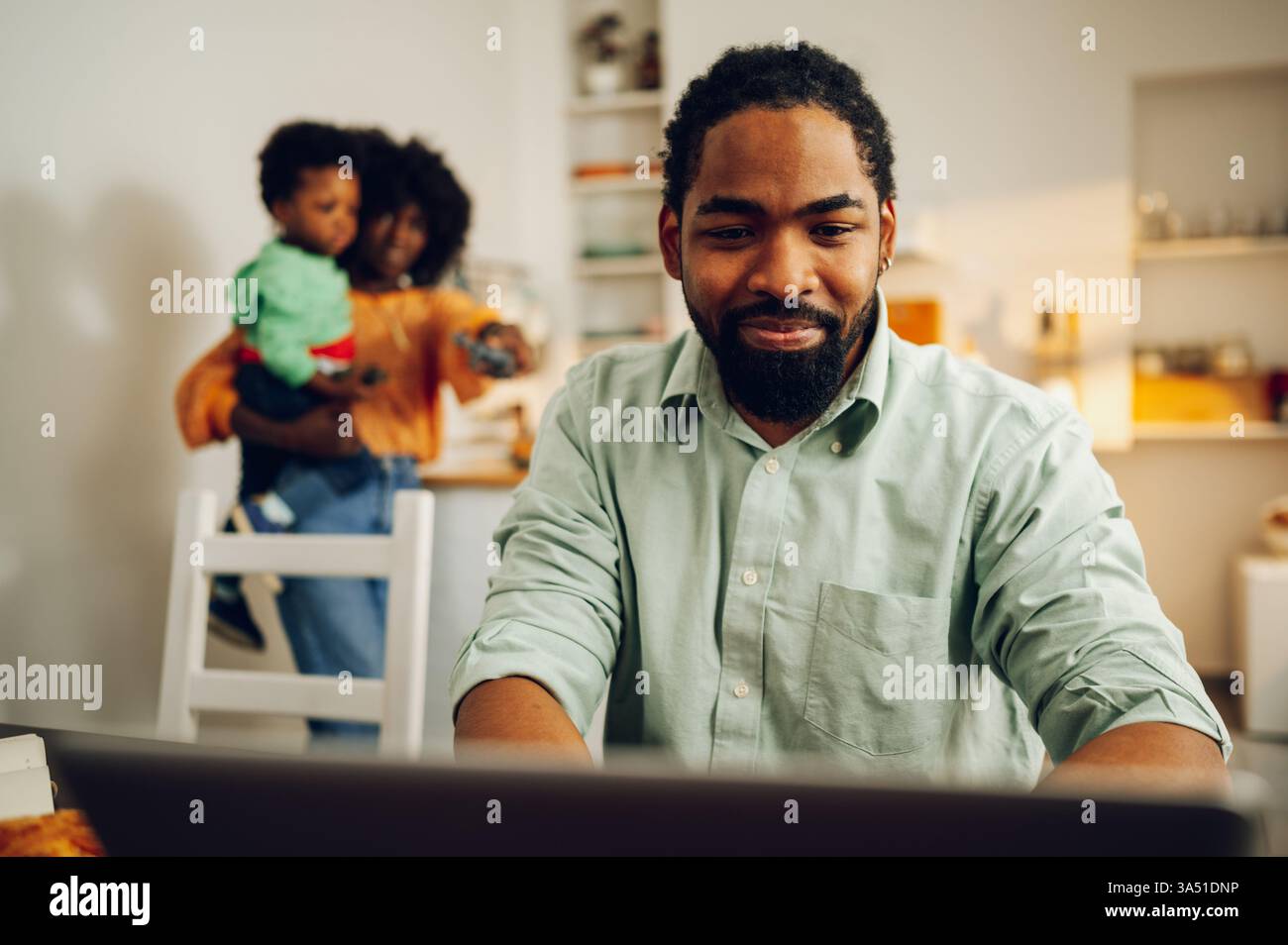 Fröhlicher multikultureller Familienmoment mit einem lässigen Geschäftsmann, der zu Hause an einem Laptop arbeitet, während seine Frau und sein Sohn in der Küche stehen. Eine entspannte Szene, die perfekt für Fernarbeit, Familienleben und Lifestyle-Kommunikation geeignet ist. Geeignet für Home Office, Technologie oder multirassische Familienkampagnen. Stockfoto